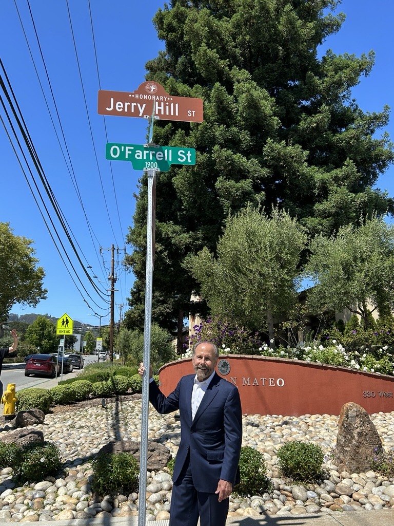 A man in a dark suit stands beside a street sign pole bearing honorary and standard street signs.