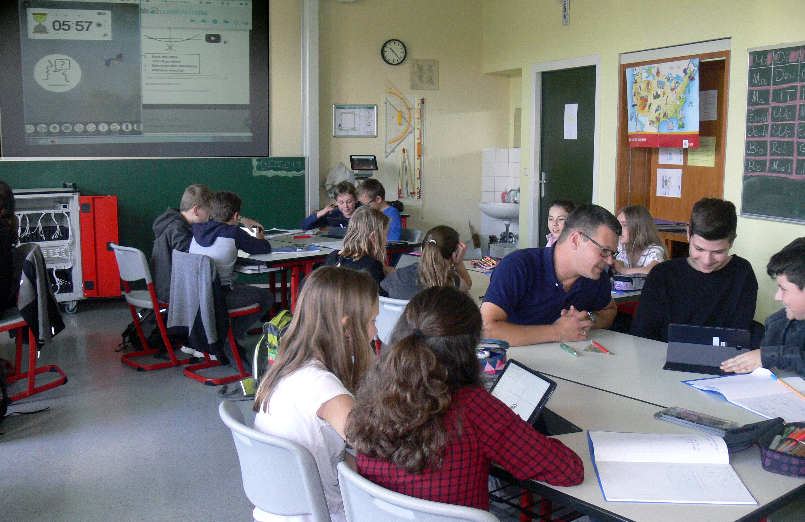 Wide classroom view showing students using tablets and a teacher seated among them.
