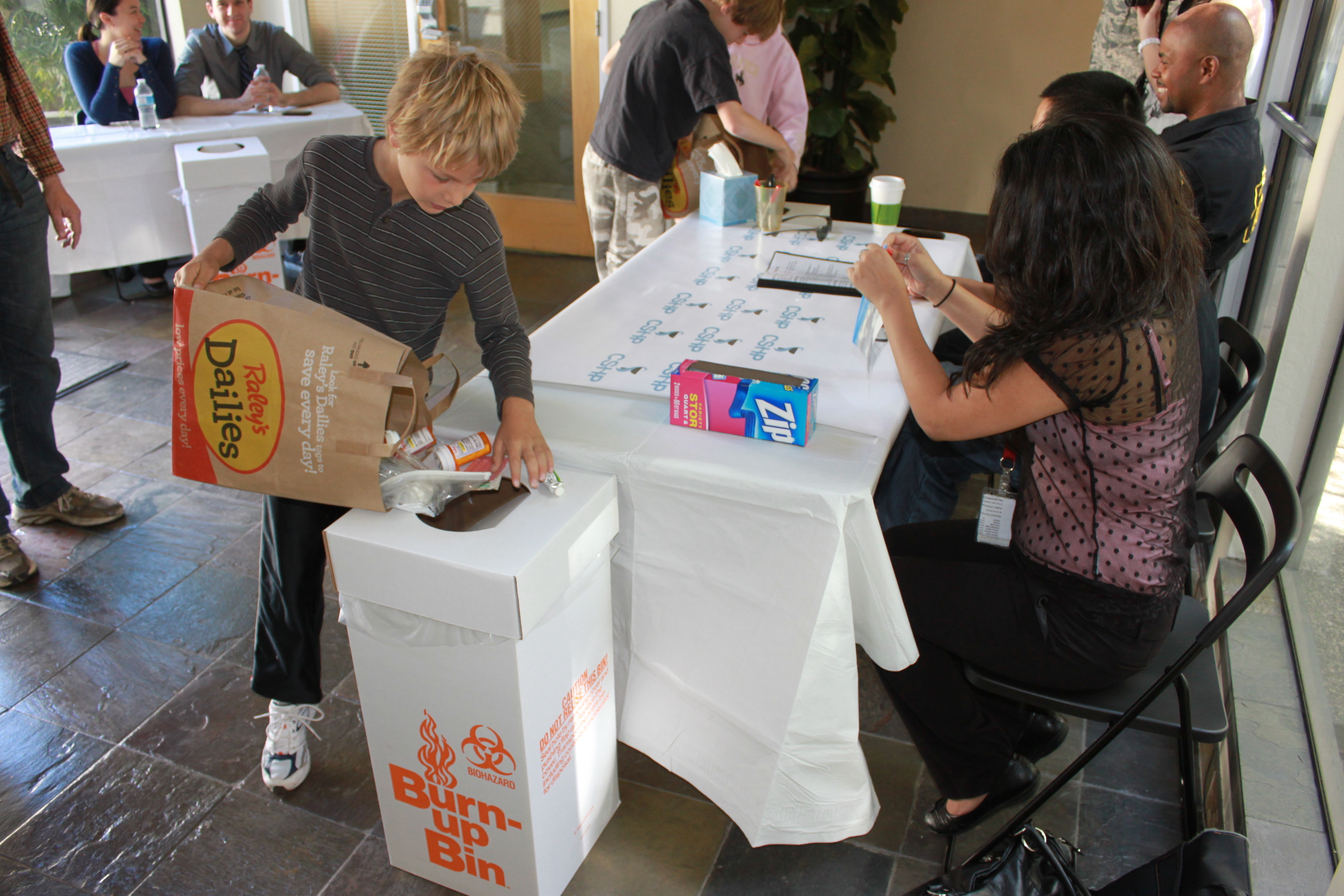 A child deposits a bag of medications into a white biohazard drop box at an outdoor event.