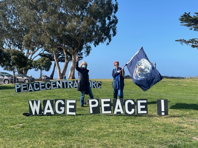 Two people hold large signs reading 'Wage Peace!' at an outdoor park on a sunny day.