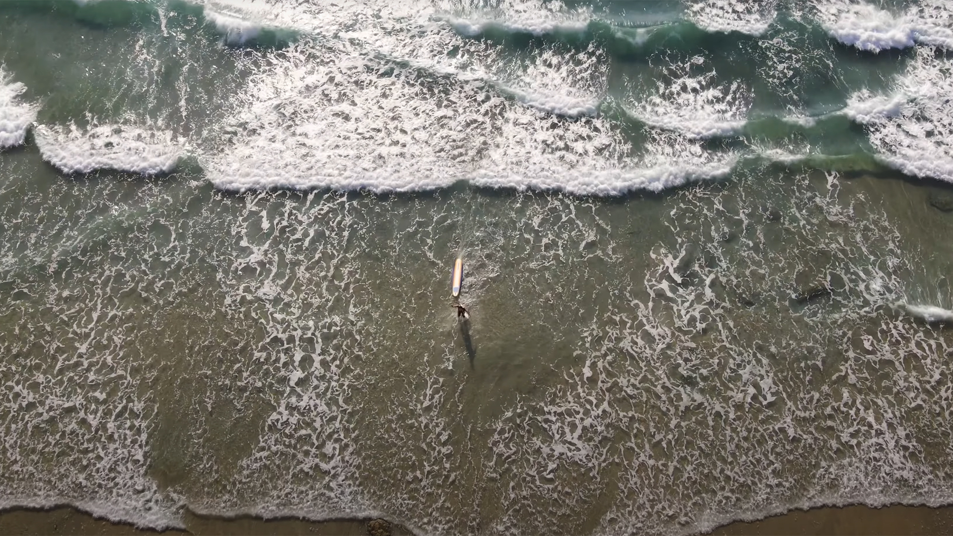 Aerial view of a surfer on a board in shallow breaking surf near shore.