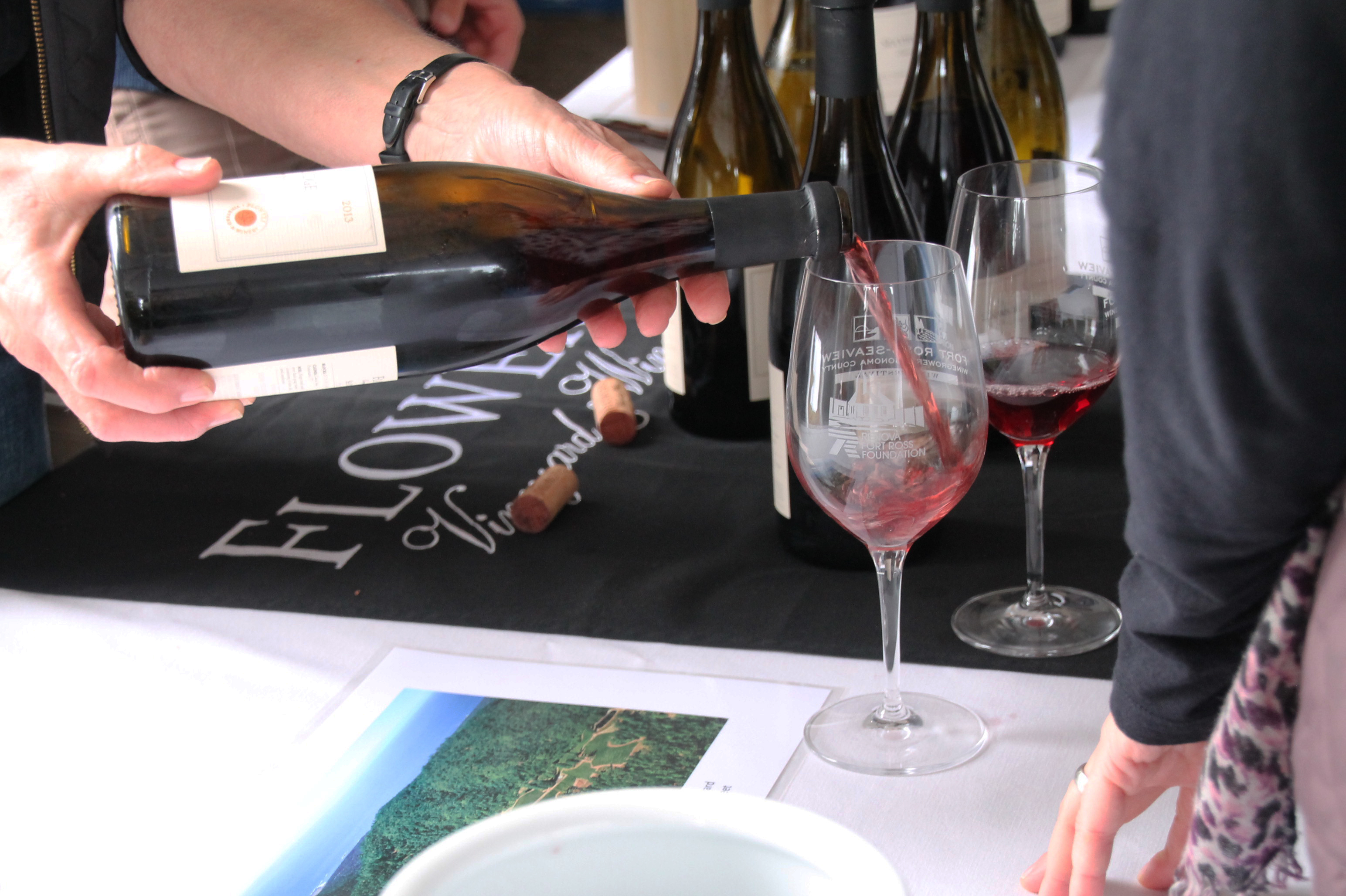 A person pouring red wine from a bottle into a stemmed glass at a tasting table.