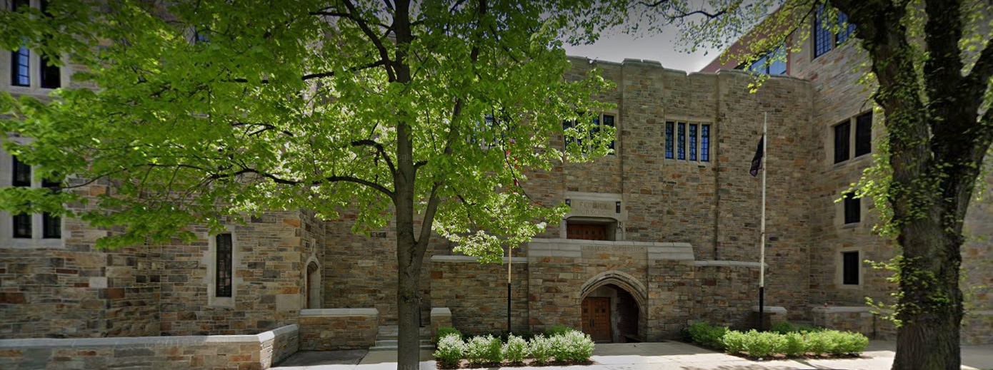 Gothic stone school building with arched entryway, flagpole, and mature trees.