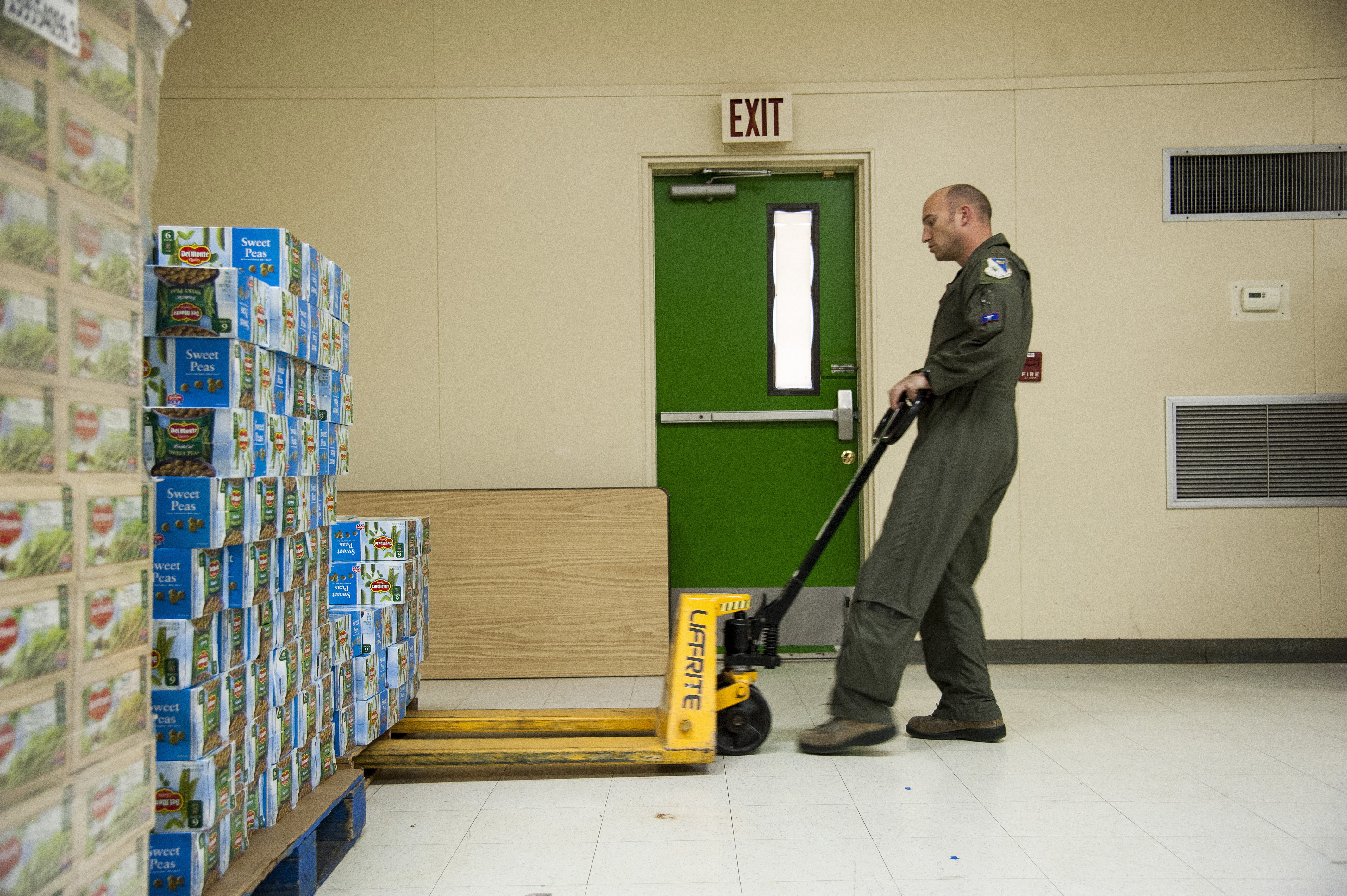 A person in a flight suit using a pallet jack to move canned goods in a food pantry warehouse.