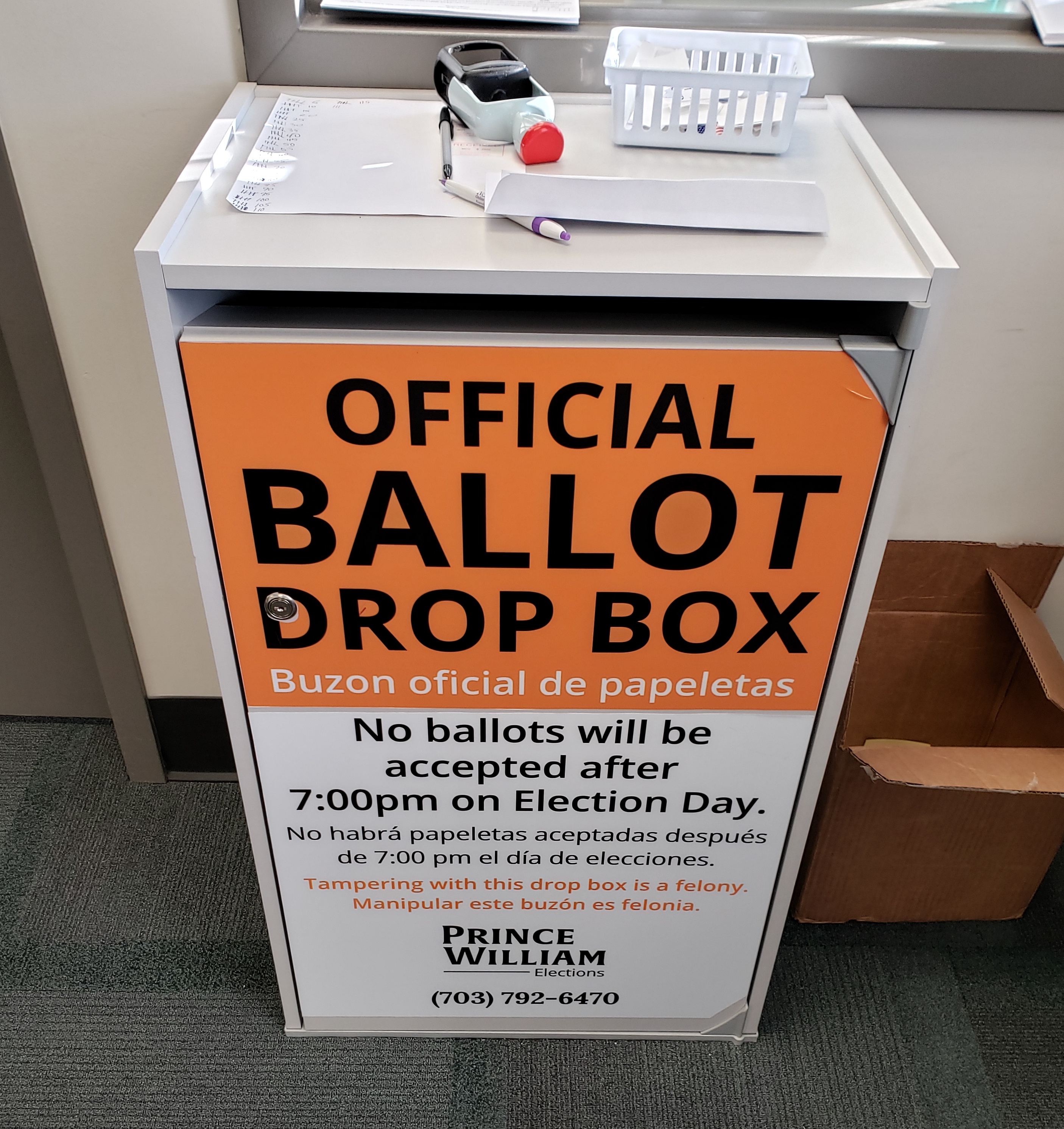 A white official ballot drop box with orange bilingual signage on a carpeted floor.
