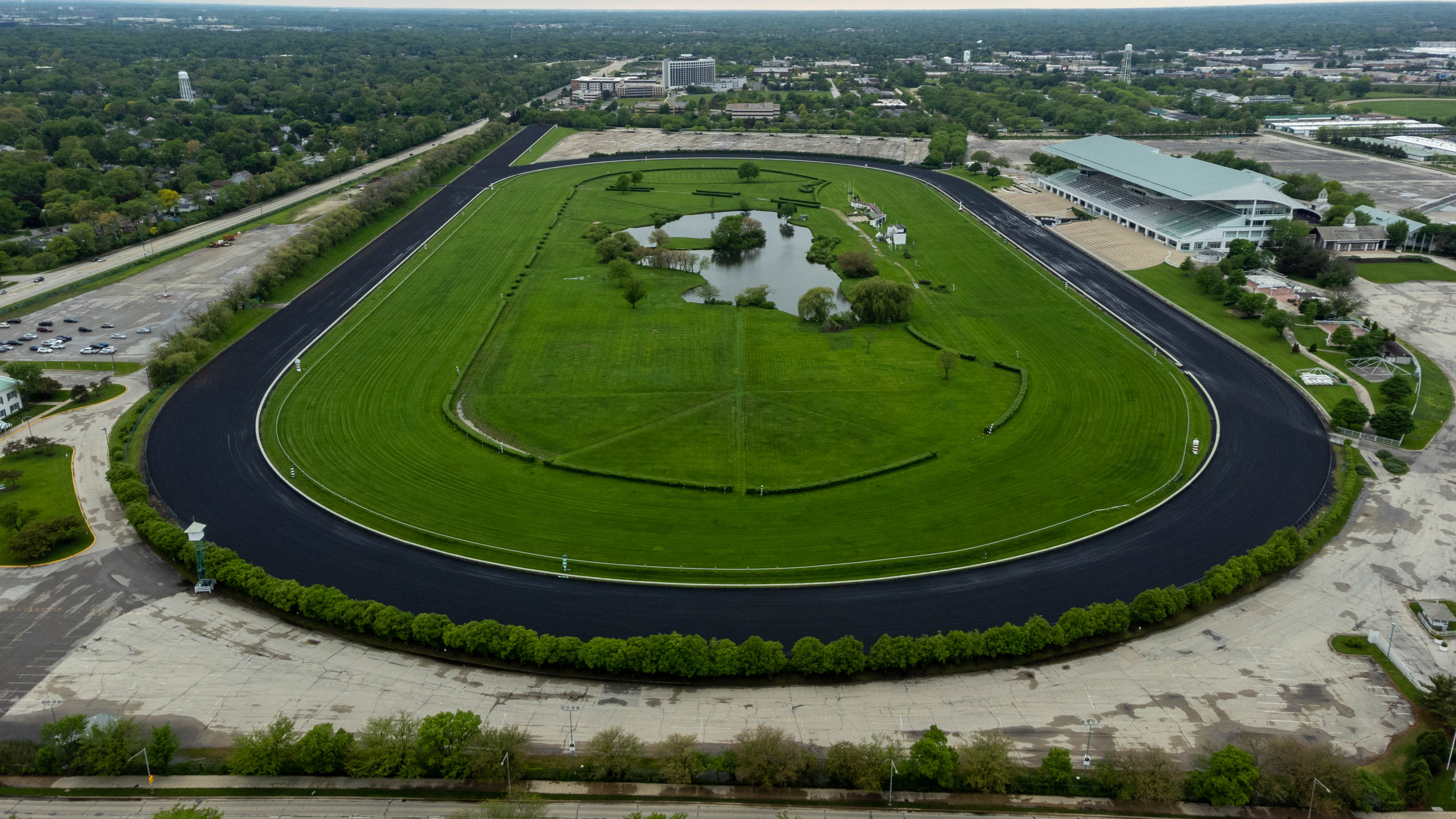 Aerial view of an oval horse racing track with green infield, pond, and suburban surroundings.