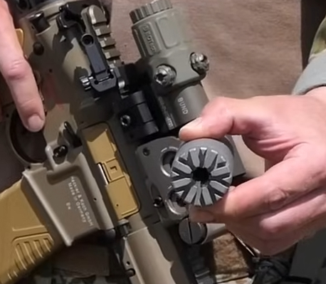Hands holding a cylindrical rifle suppressor next to a tan rifle with optics.