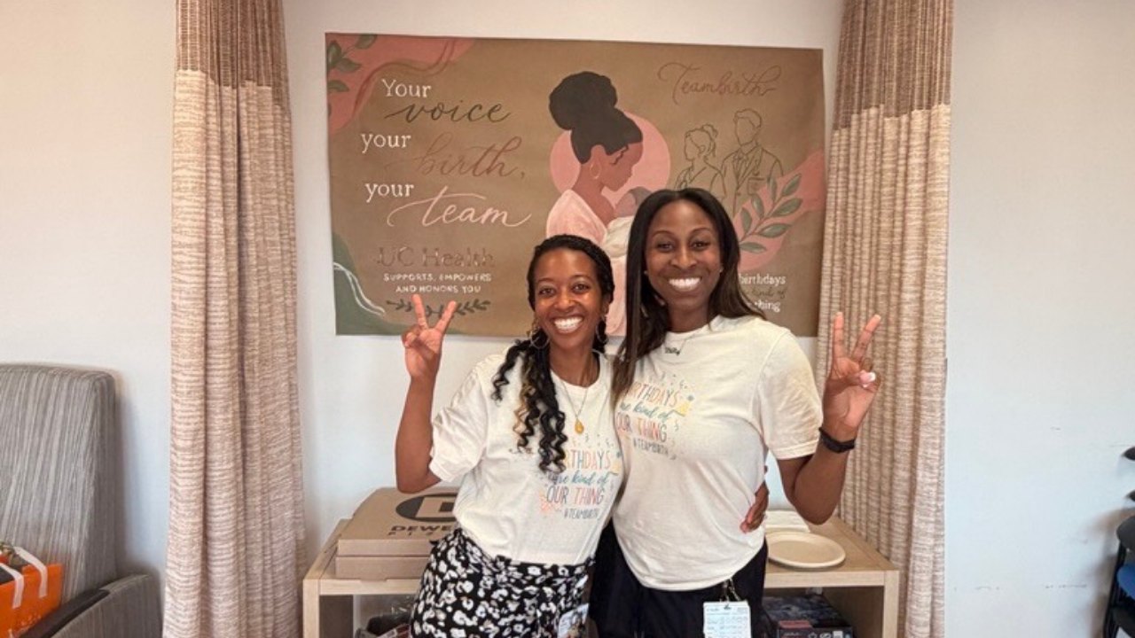 Two women in white T-shirts pose in front of a wall sign reading 'Your voice your birth your team.'
