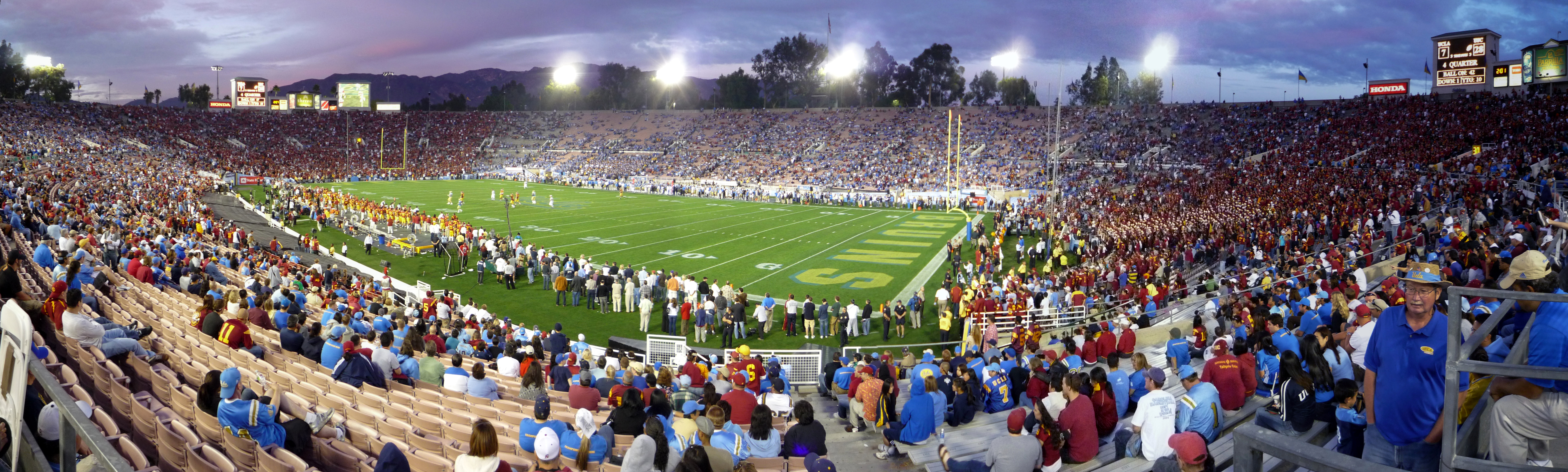 Panoramic view of the Rose Bowl stadium lit by floodlights at dusk, stands filled with fans.