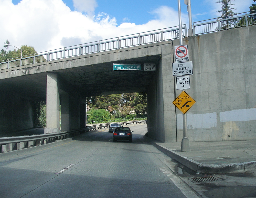 Westbound view through a concrete underpass with road signs and a sedan ahead.