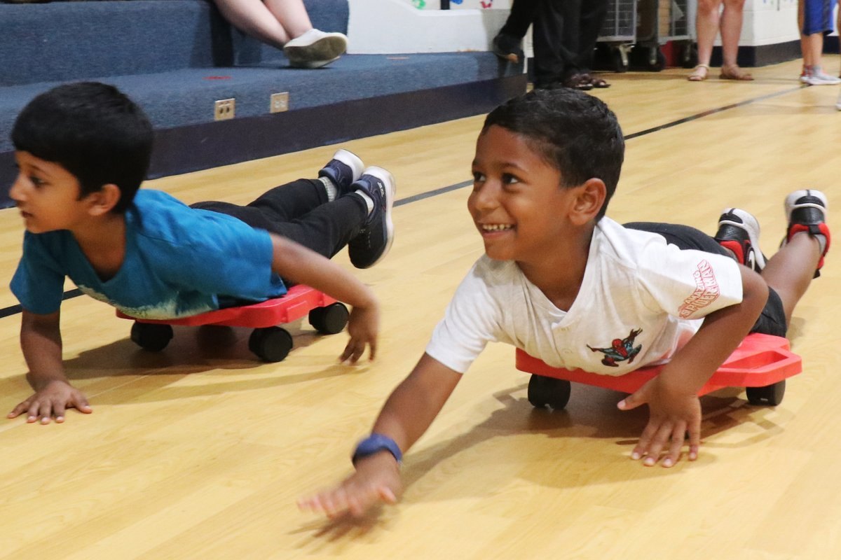 Two young children lying on red scooter boards on a gymnasium floor, smiling.