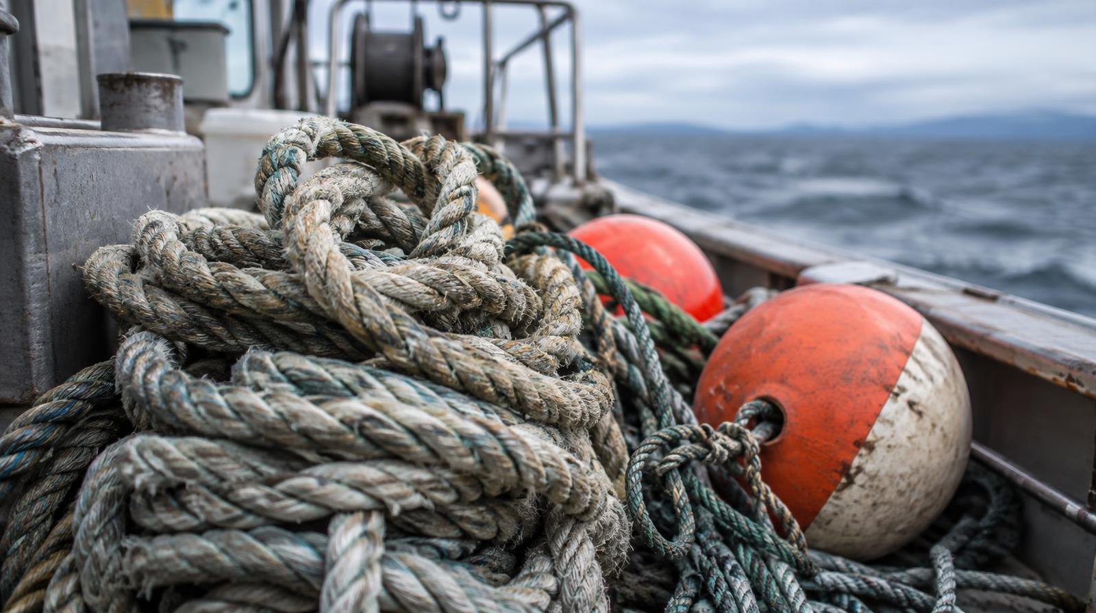 A coil of thick rope and a buoy line tangled together on a boat deck with ocean in the background.