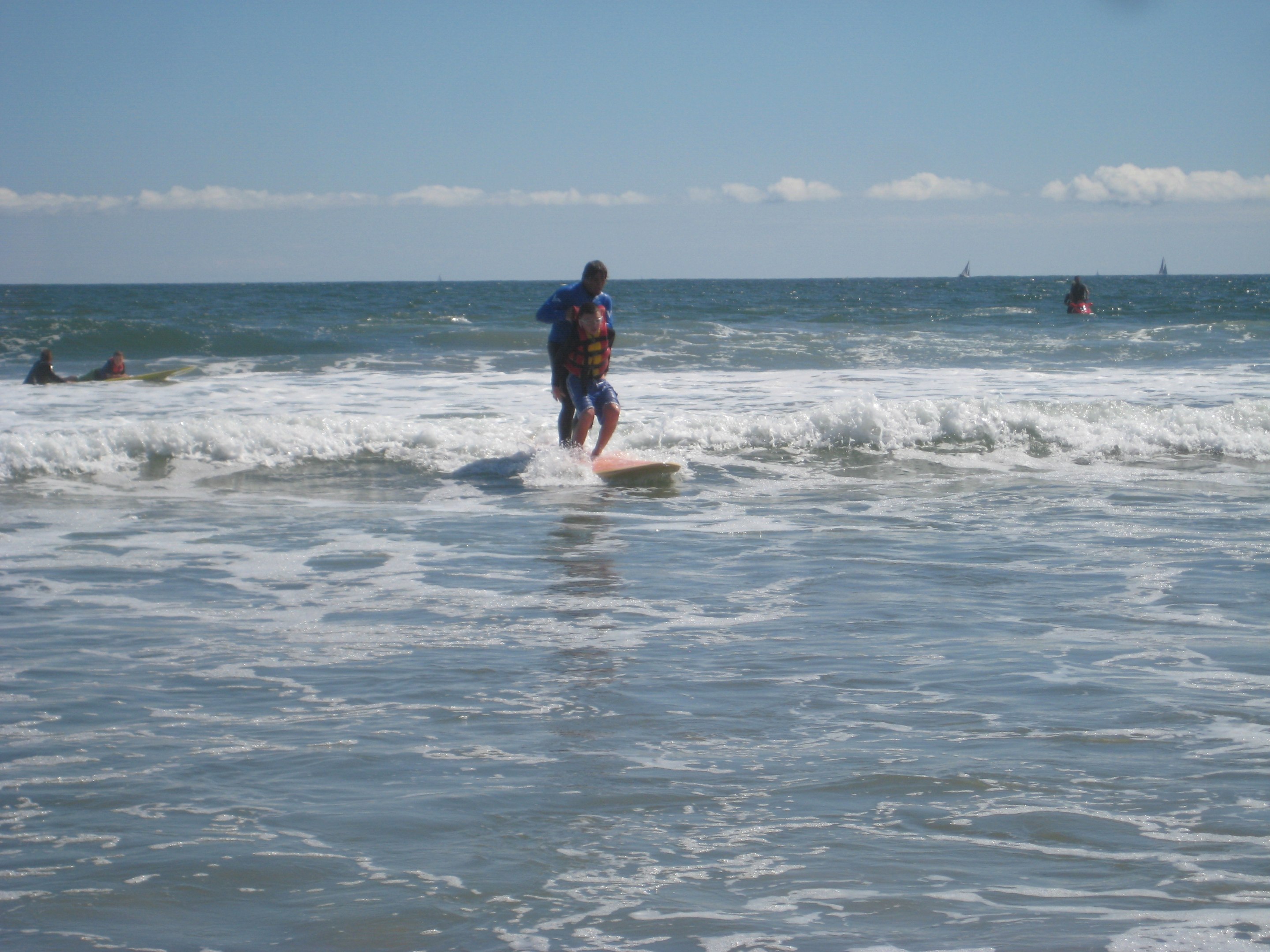 A person on a surfboard in shallow surf, guided by an assistant standing behind them.