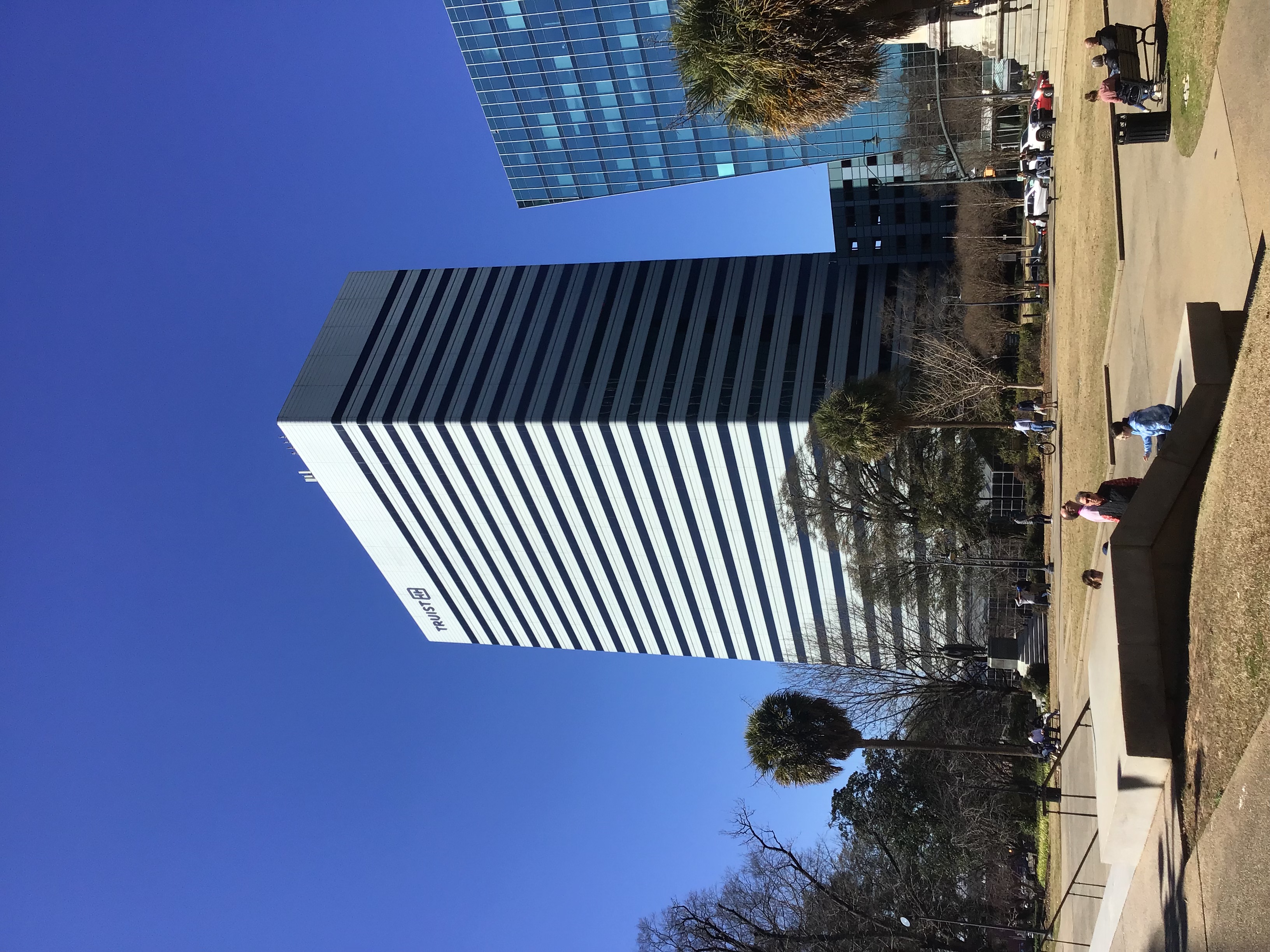 Tall glass office tower seen from a sunny public plaza with palm trees.