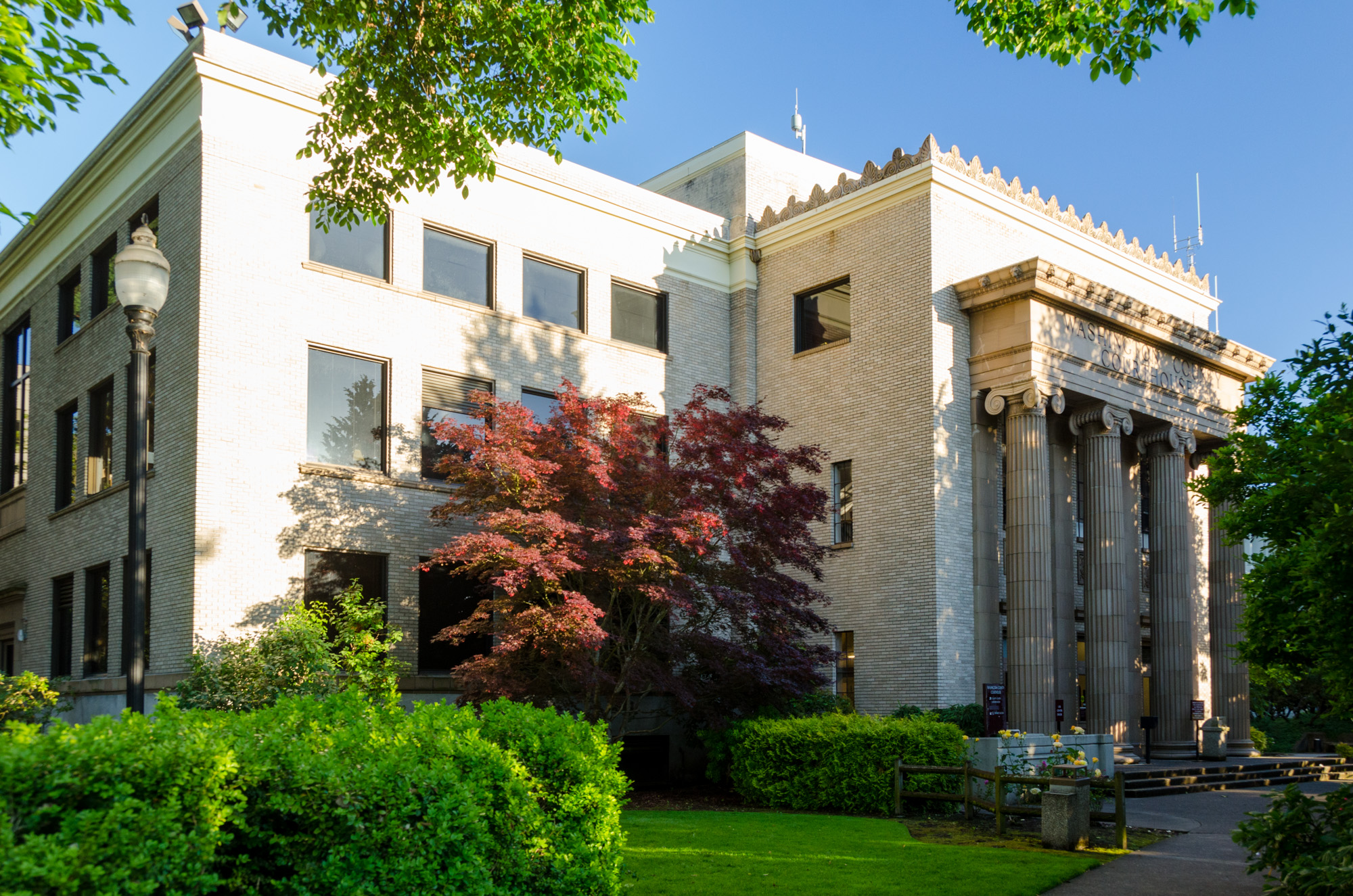 Neoclassical courthouse building with tall columns, red ornamental shrub, and green trees in front.