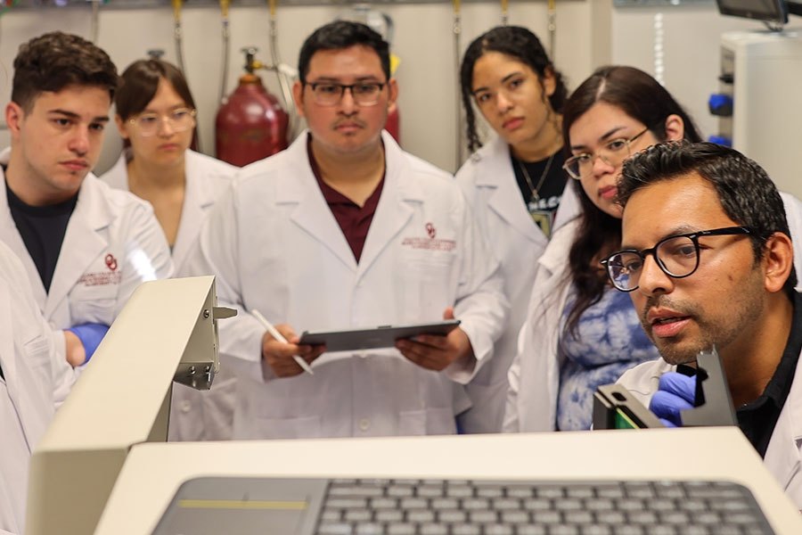 Six people in white lab coats observe a demonstration around laboratory equipment.