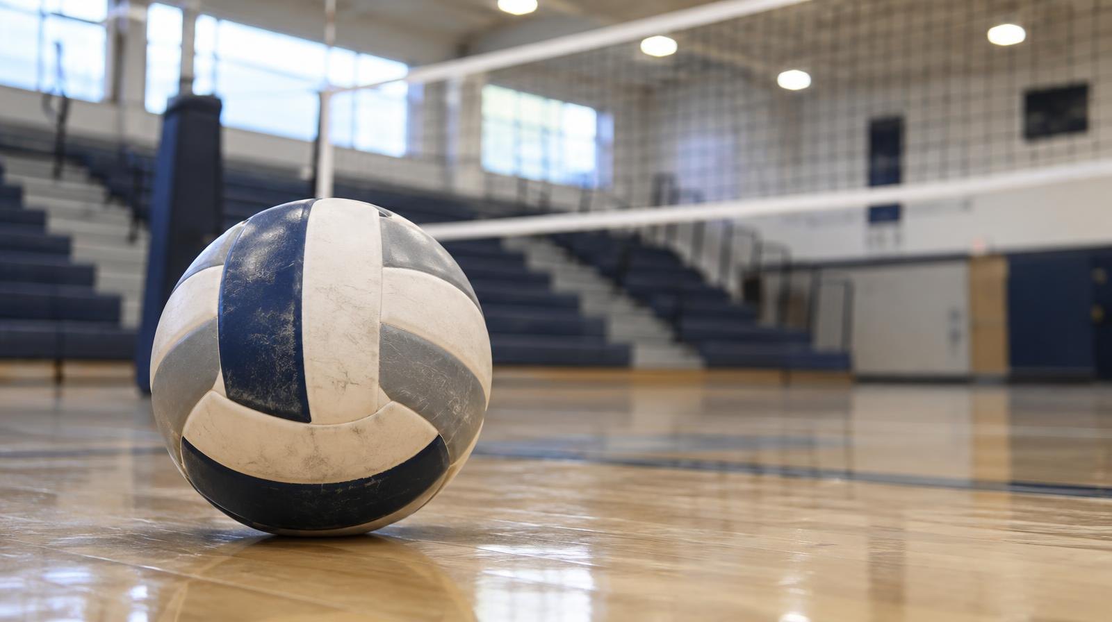 A volleyball resting on a hardwood gymnasium floor near a net with bleachers in the background.