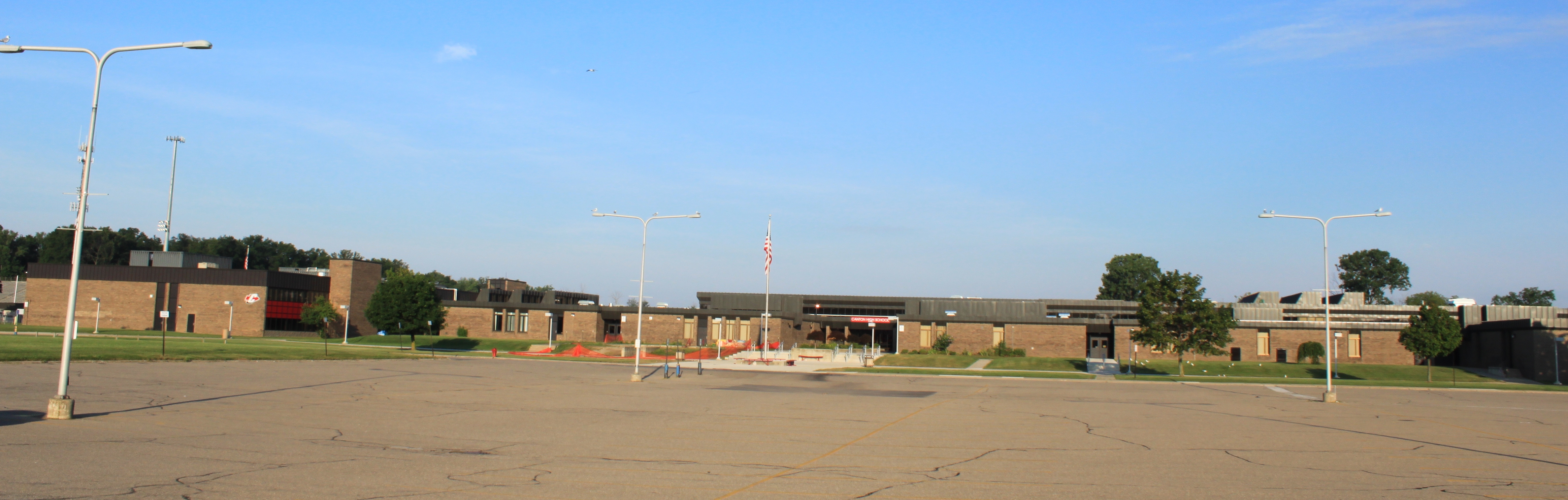 Wide single-story brick school building with a large parking lot and flagpole.