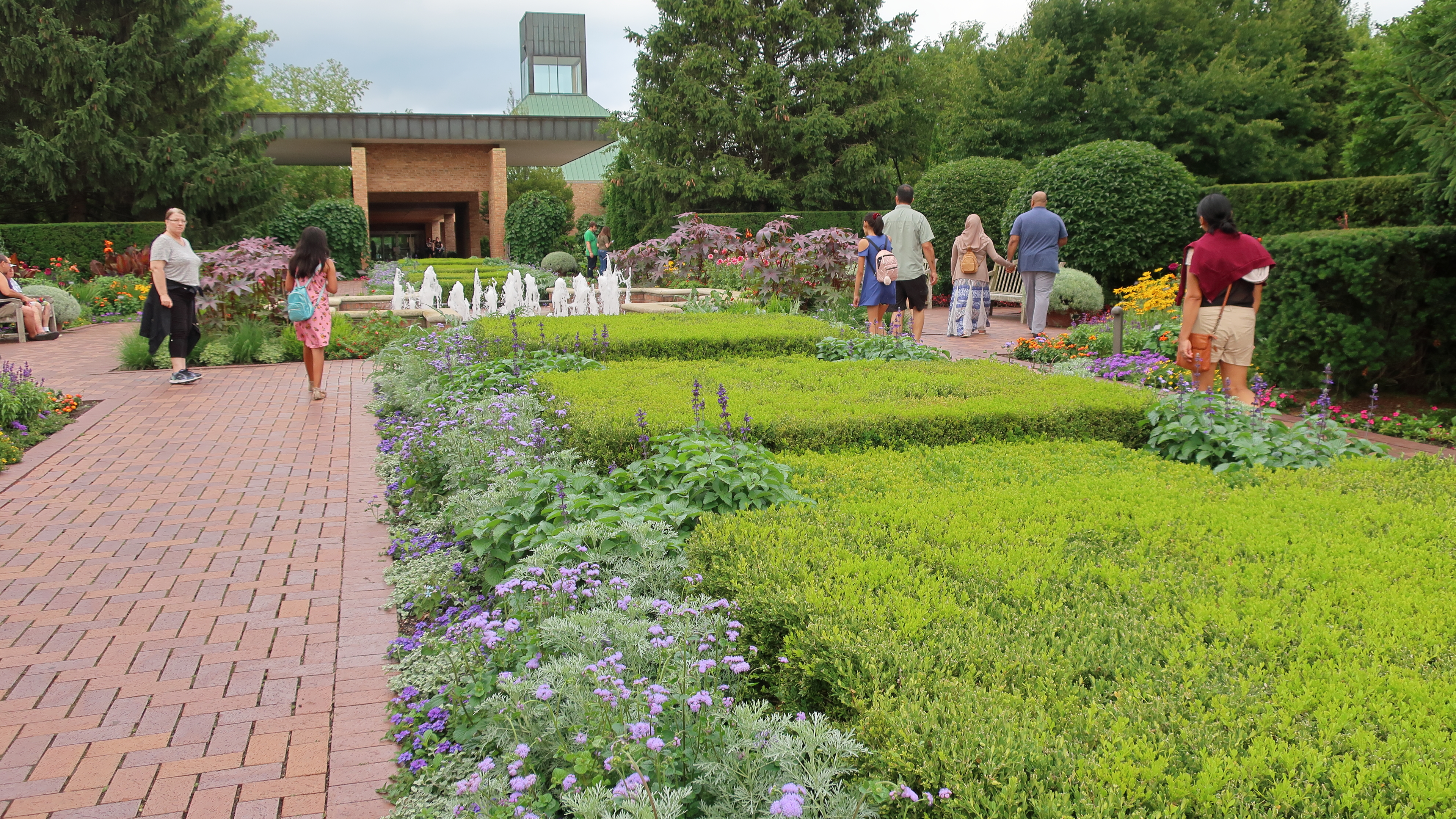 Visitors walking along brick paths through a formal garden with trimmed hedges and flowering plants.