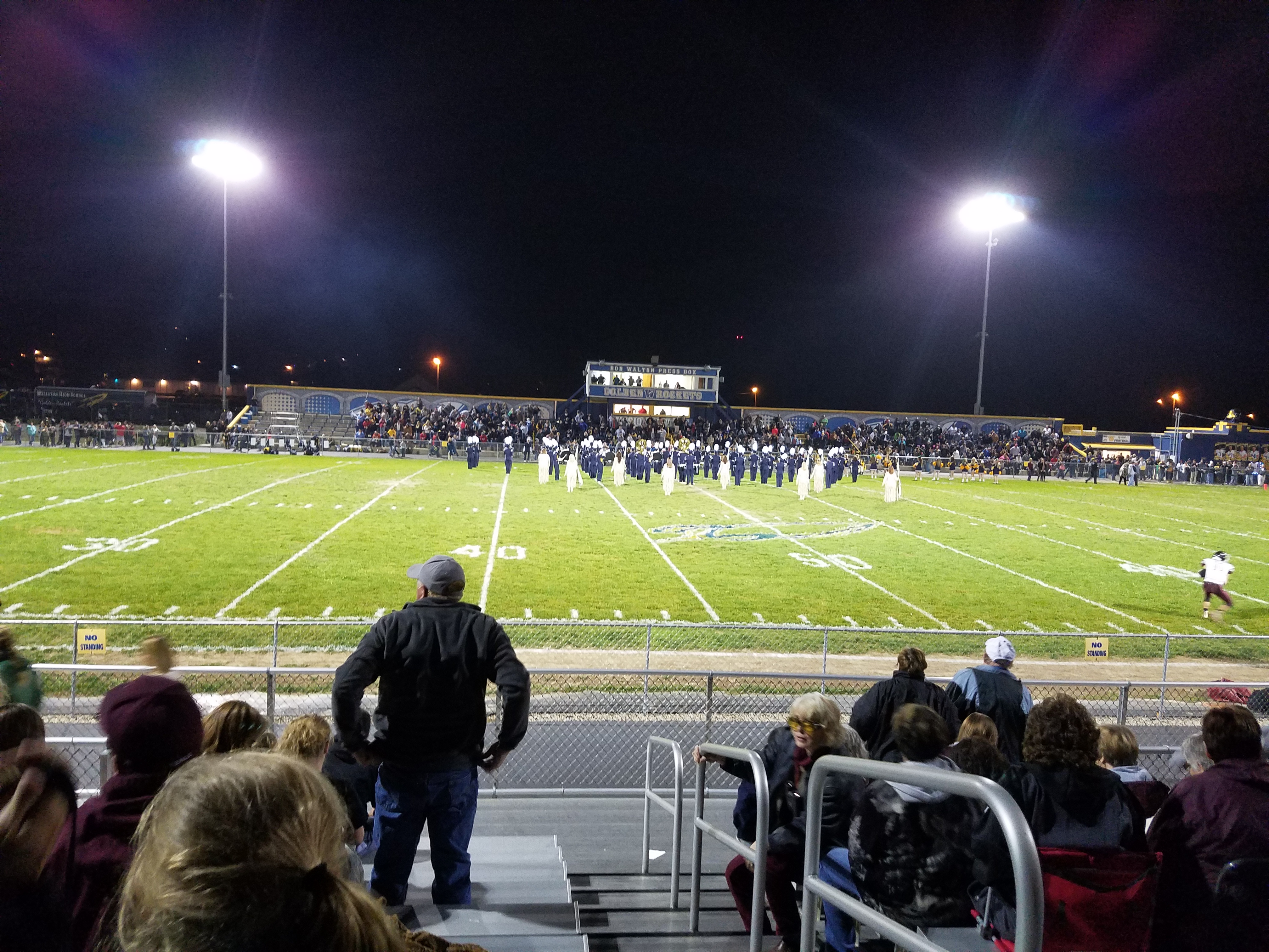 Spectators in bleachers watching a high school football game under stadium lights at night.