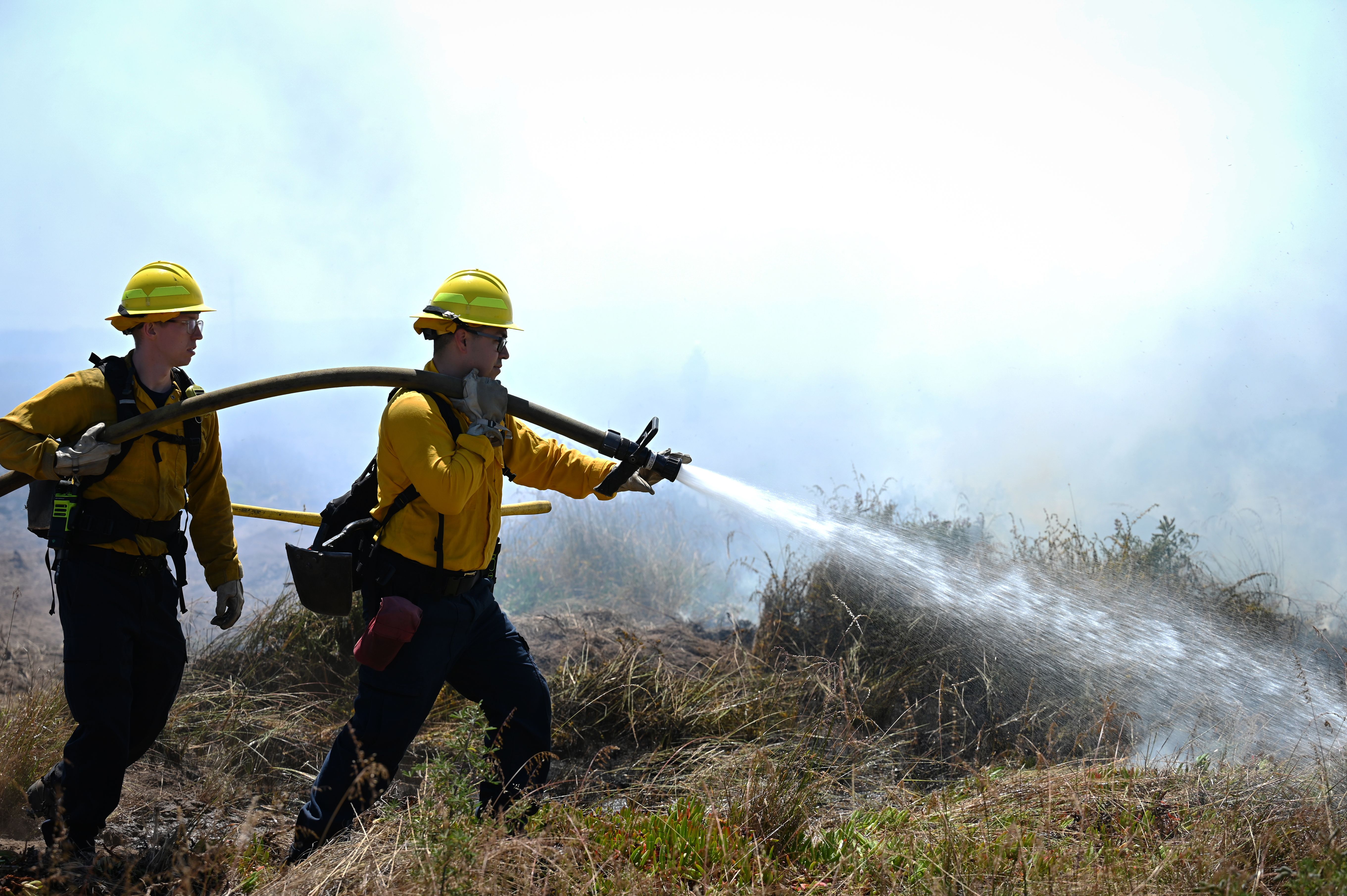 Two firefighters in yellow gear direct a hose at a prescribed burn in dry grassland.