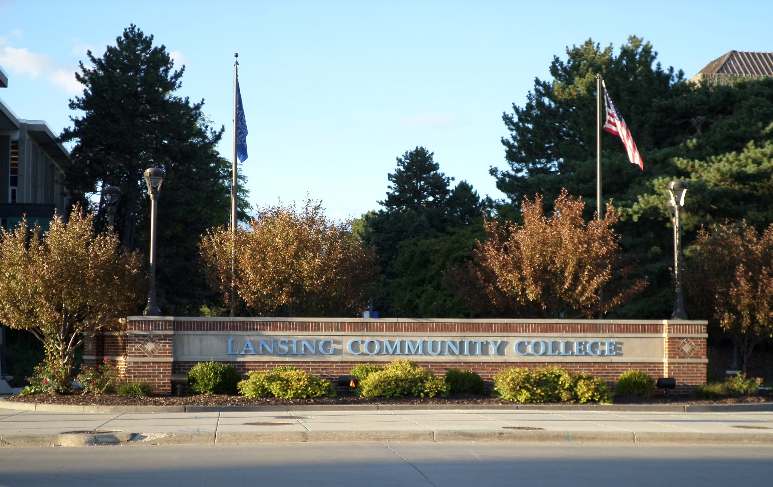 Brick entrance sign reading Lansing Community College with flagpoles and trees behind it.