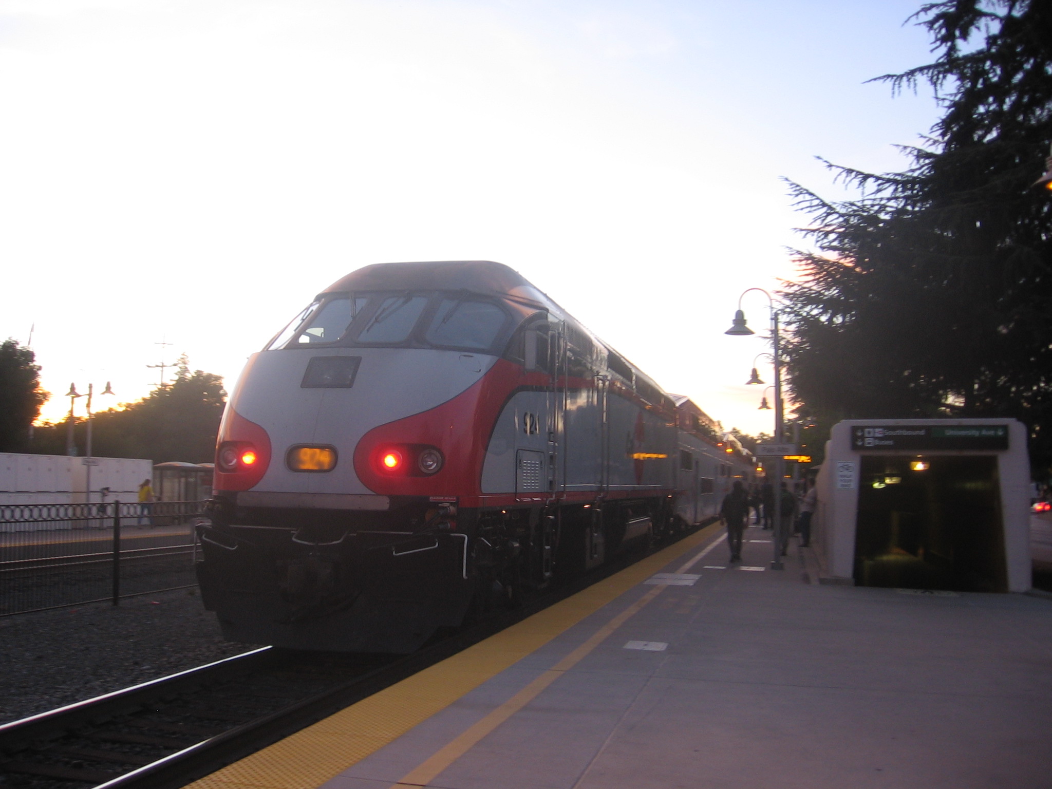 A commuter train arriving at a station platform at dusk with passengers waiting on the platform.