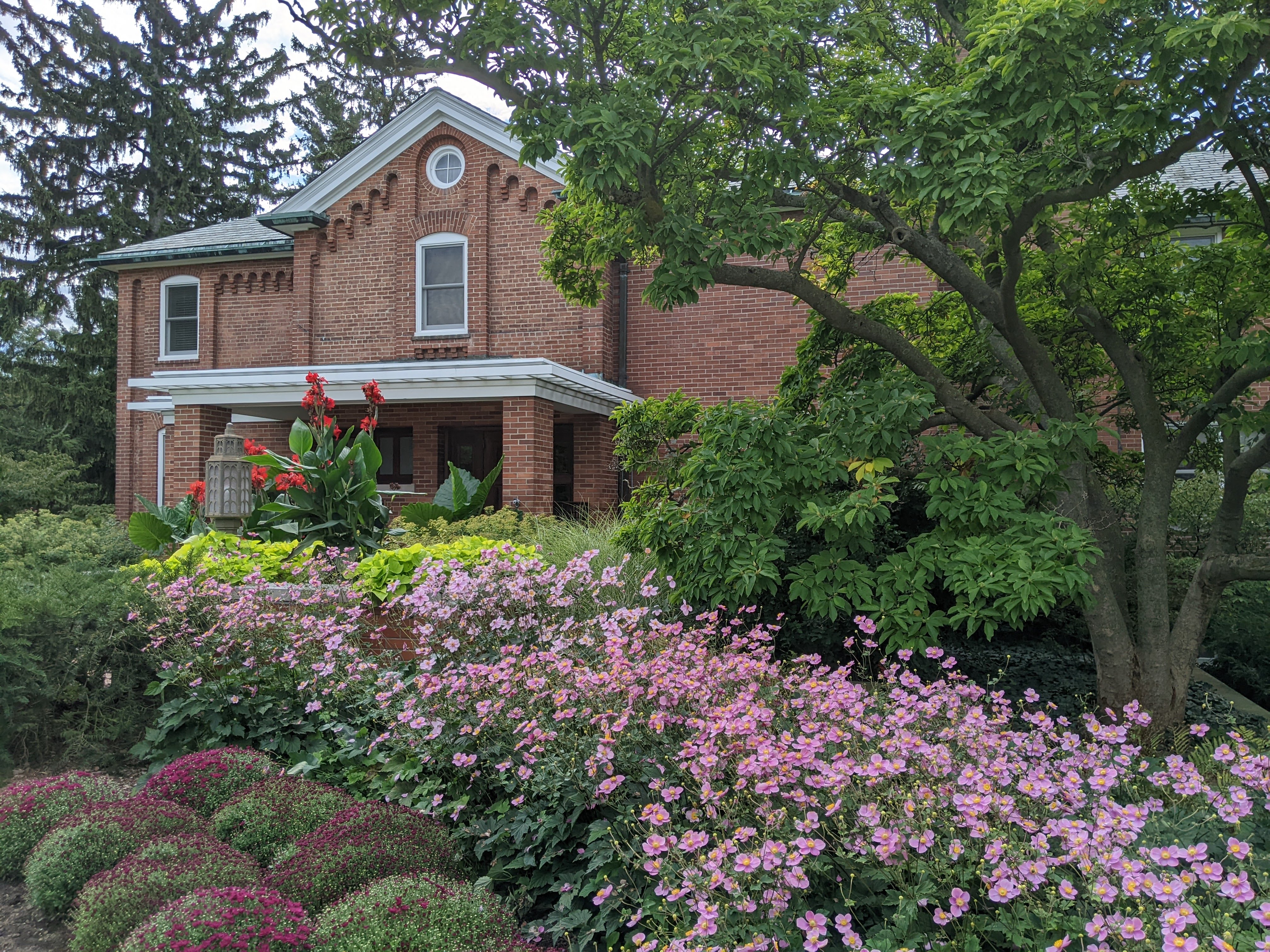 Red-brick university building behind colorful flowering gardens on a summer day.