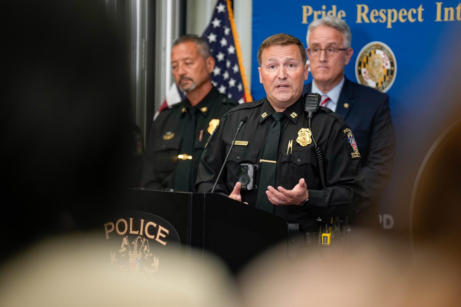 A police captain in uniform speaks at a podium labeled POLICE, flanked by two officials with an American flag behind them.
