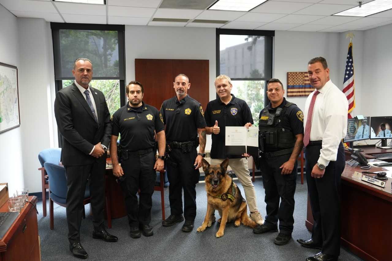 Sheriff's office personnel in uniform pose with a German Shepherd dog and a certificate indoors.