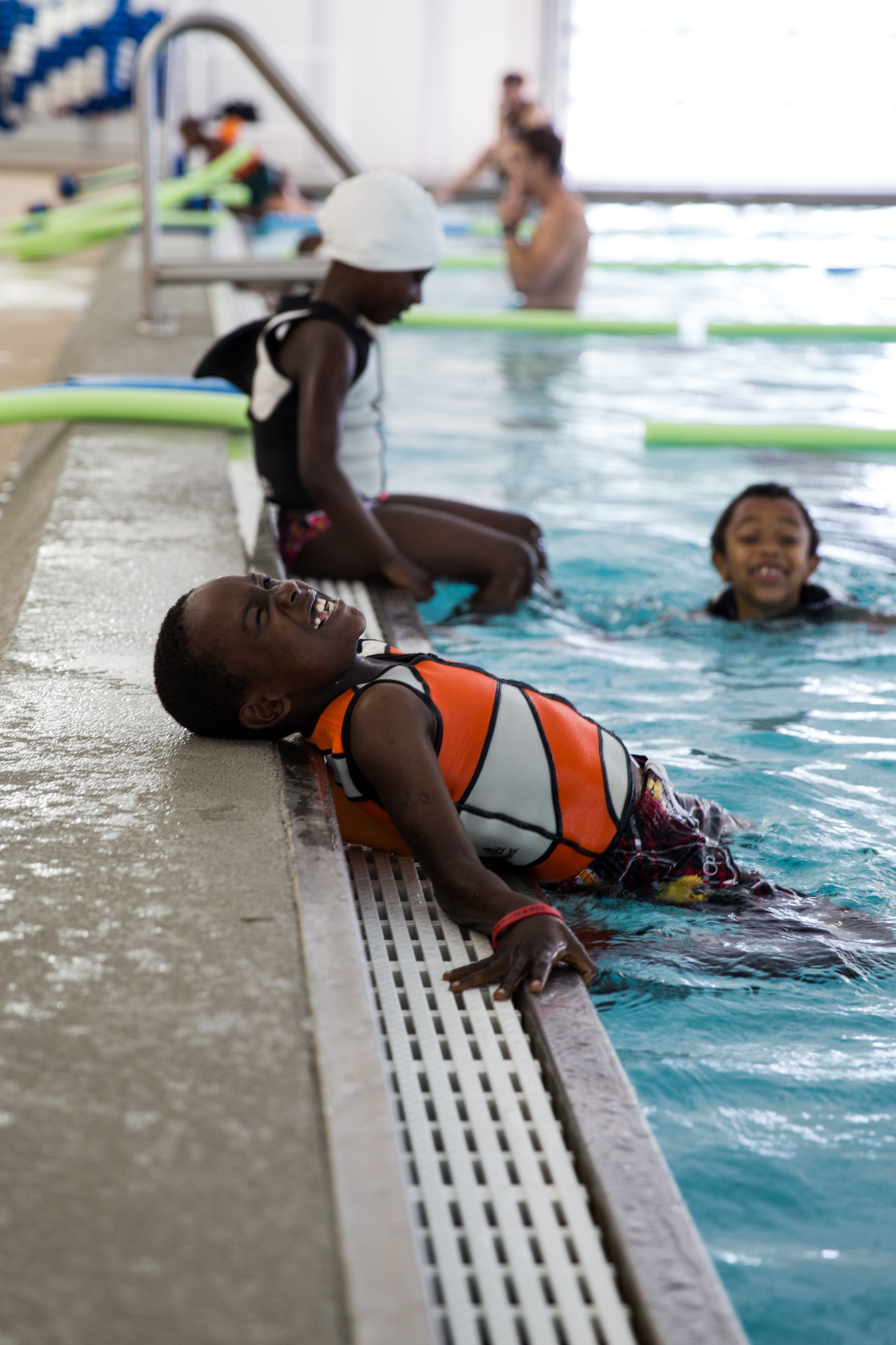 A child in an orange life vest clings to the edge of an indoor swimming pool.