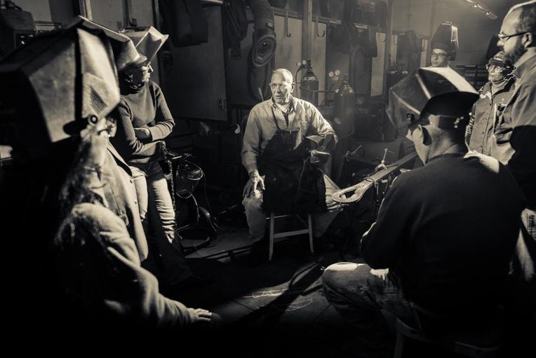 Group of people in welding helmets gathered around an instructor in an industrial workshop.