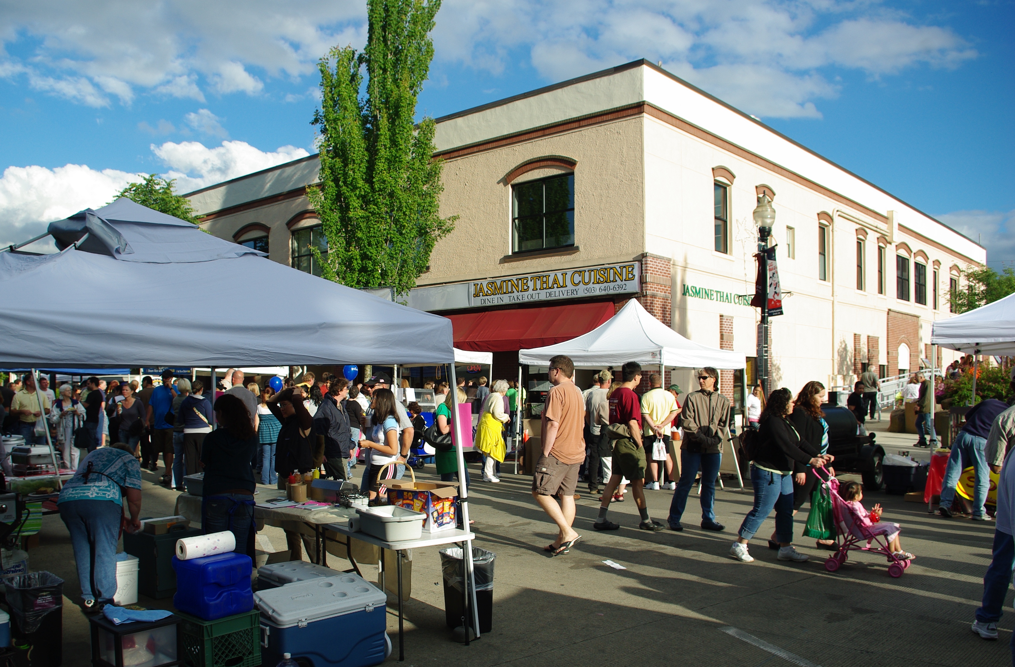 Crowds of people walking among vendor tents at an outdoor farmers market on a sunny day.