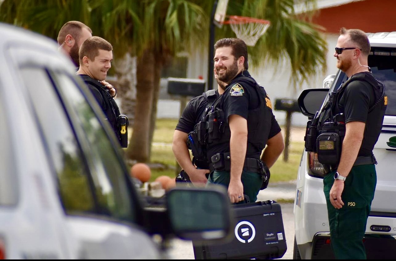 Four uniformed sheriff's deputies stand near patrol vehicles outdoors.
