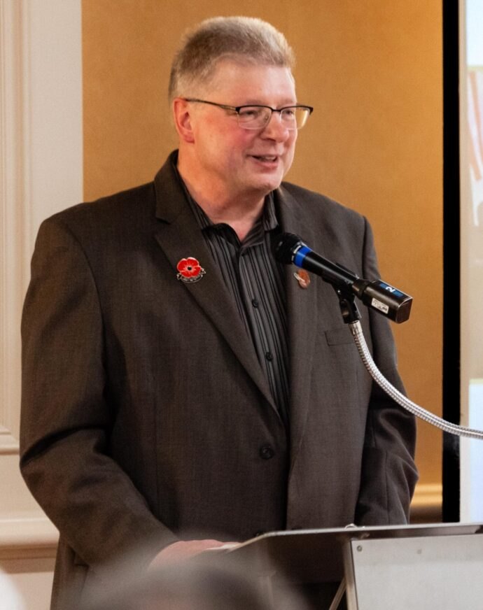 Man with glasses in a dark blazer speaks at a podium with a microphone.