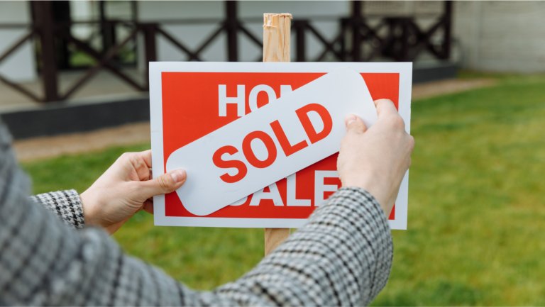 Hands attaching a red sold sign to a home-for-sale post in front of a house.