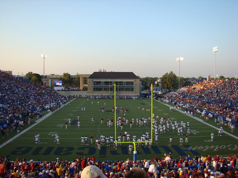 Packed football stadium with players on the field and yellow goalposts in the foreground.