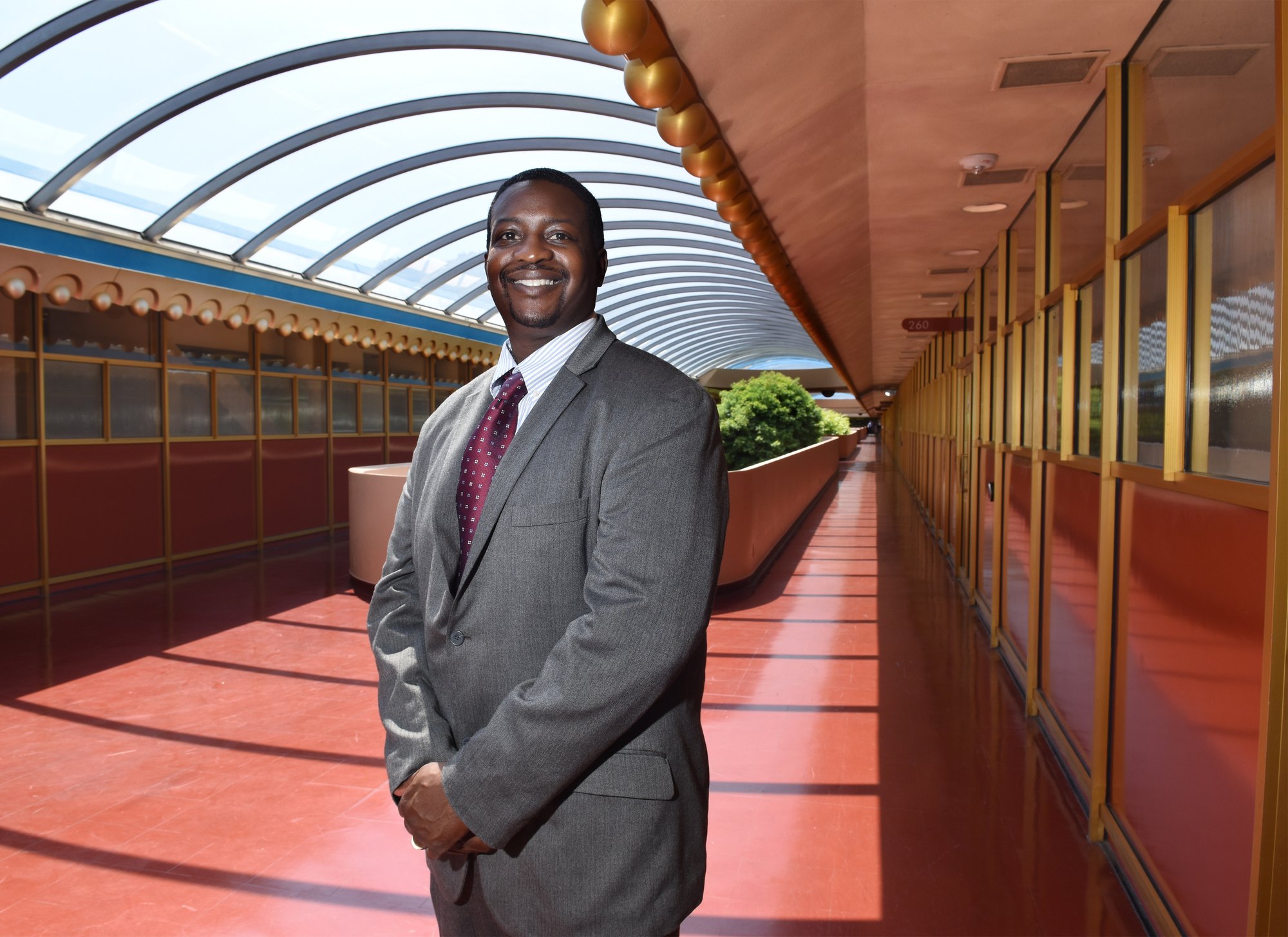 A smiling man in a gray suit and red patterned tie standing in a curved glass-roofed corridor.