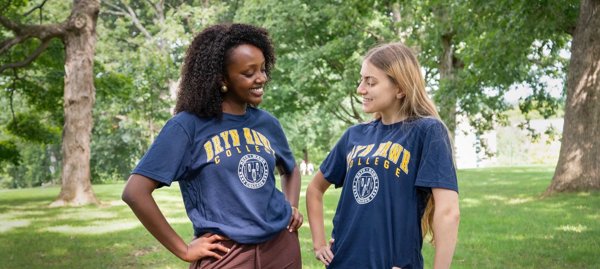 Two smiling students wearing navy Bryn Mawr College t-shirts stand outdoors on campus.