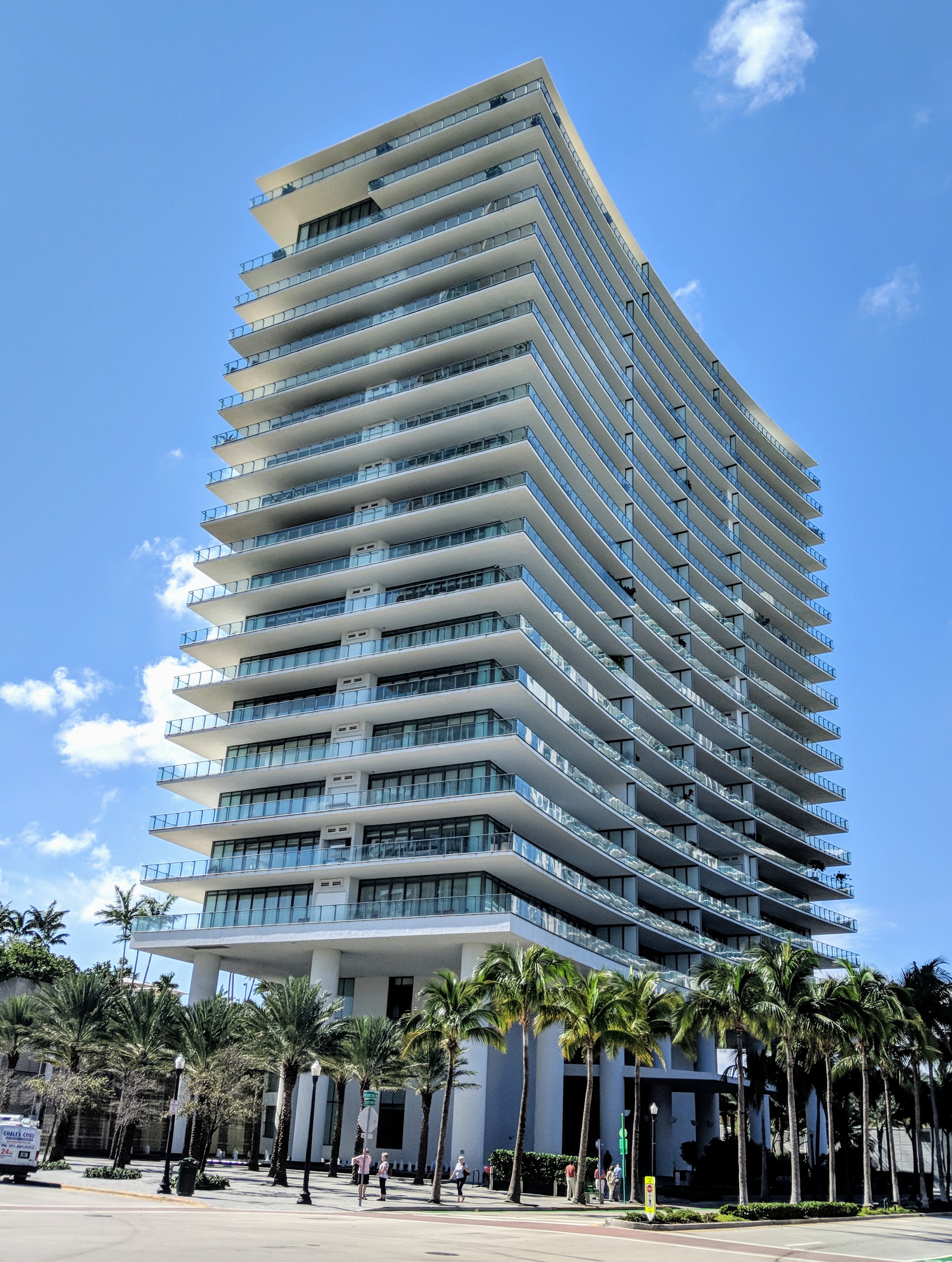 Tall white condominium tower with glass balconies and palm trees lining the street.