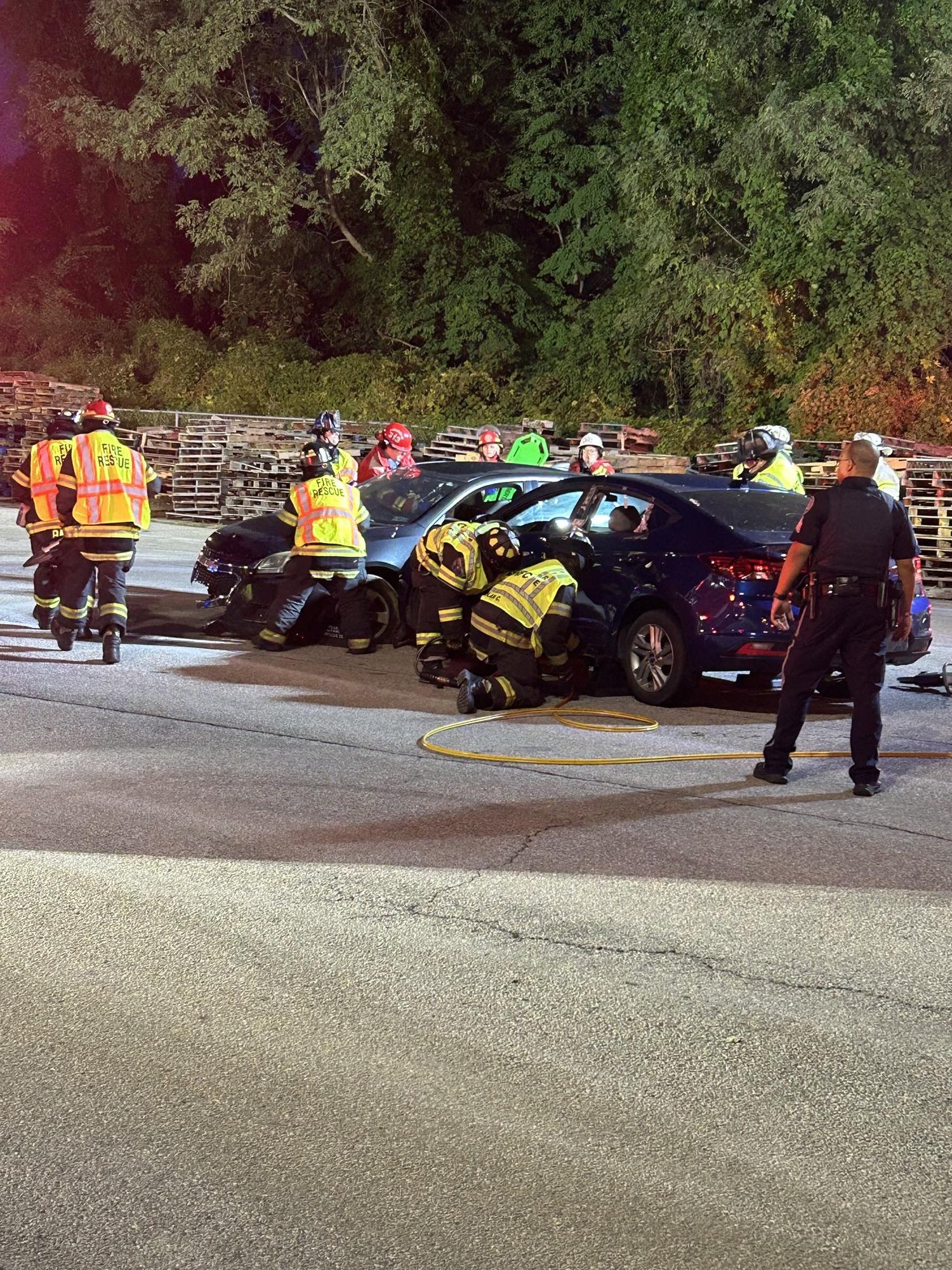 Firefighters in high-visibility vests work around a heavily damaged vehicle at night with hoses on the pavement.