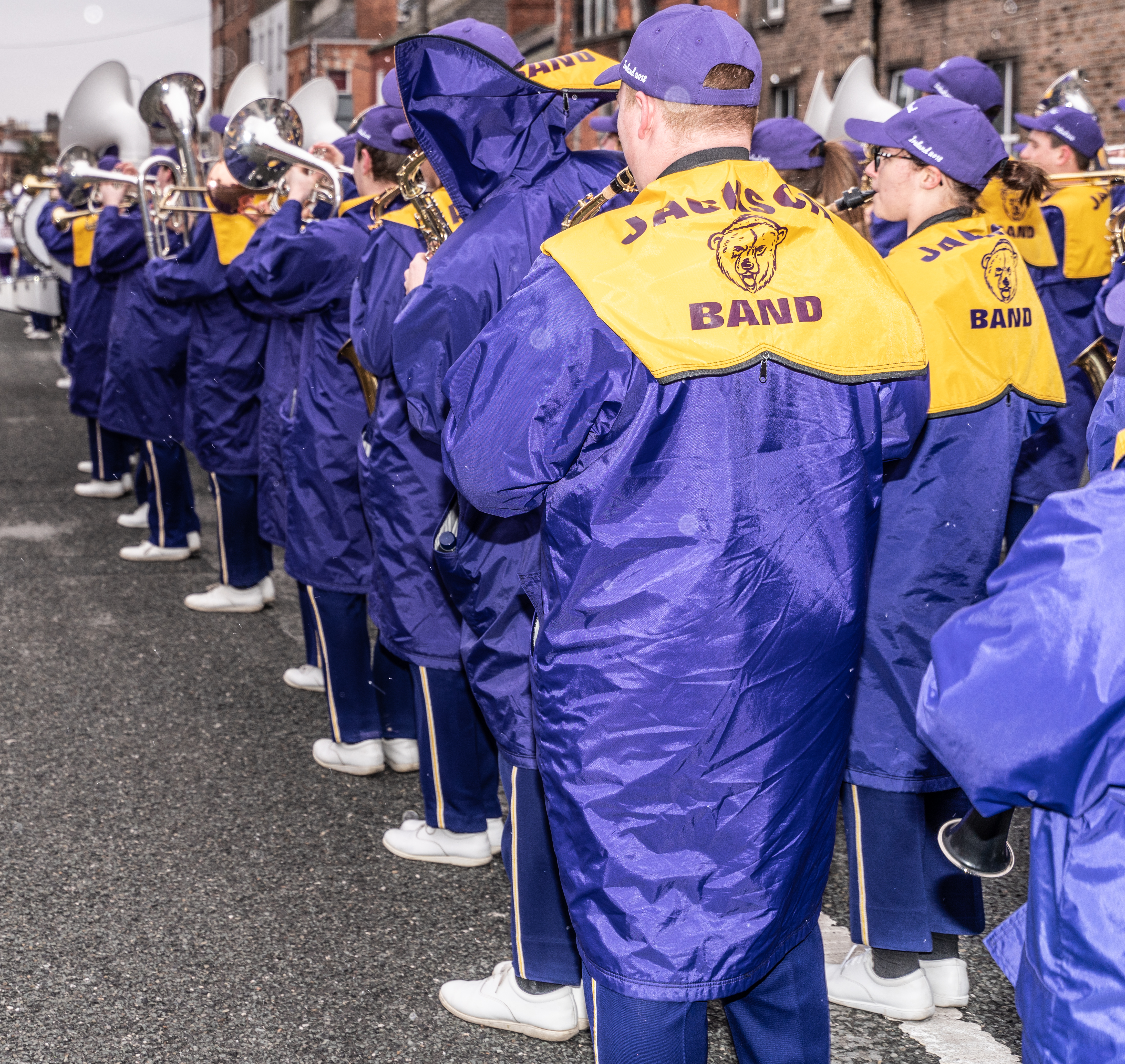 High school band members in matching jackets stand in formation on a street.