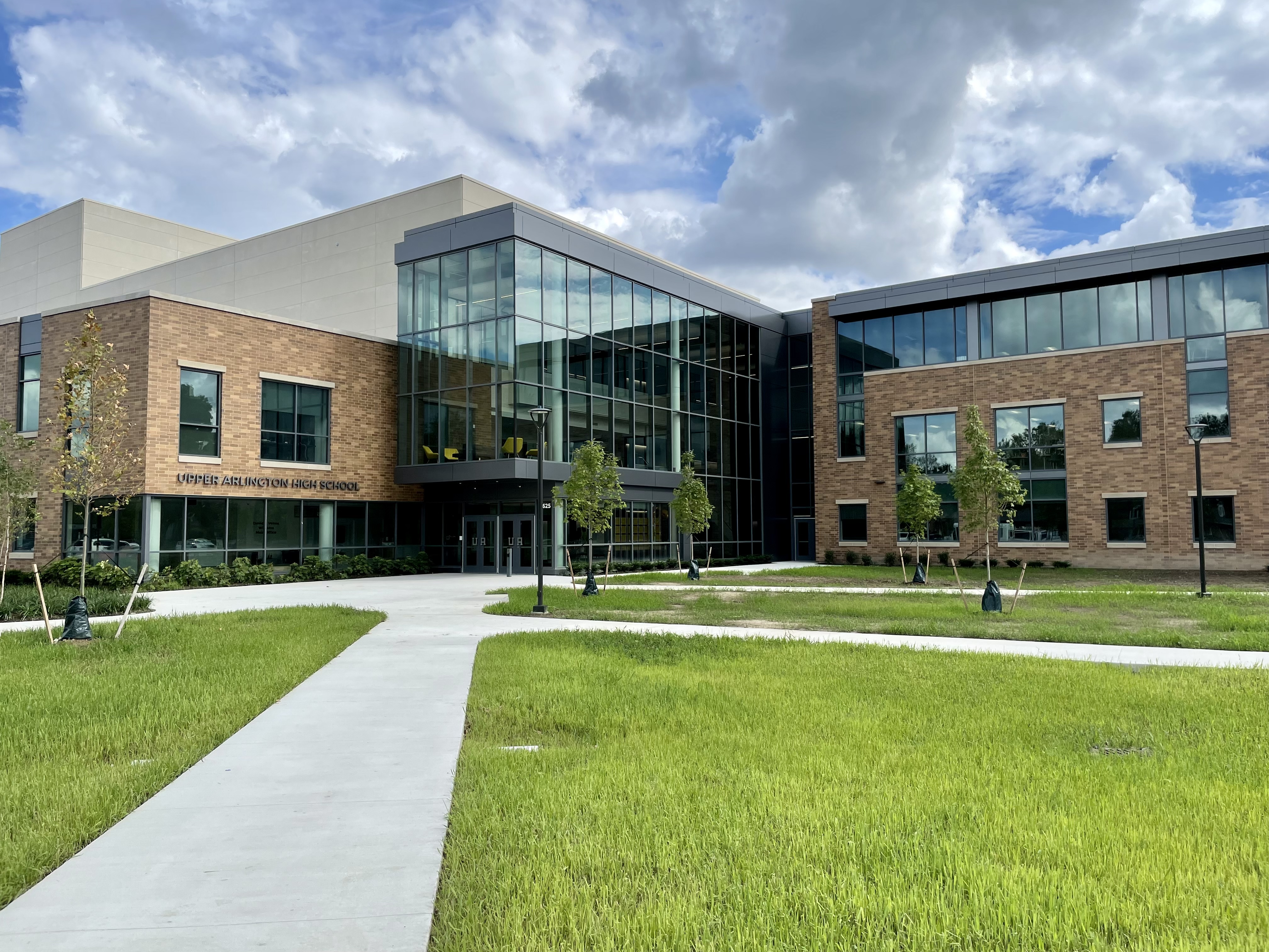 Modern brick and glass school building exterior with landscaped walkway under partly cloudy sky.
