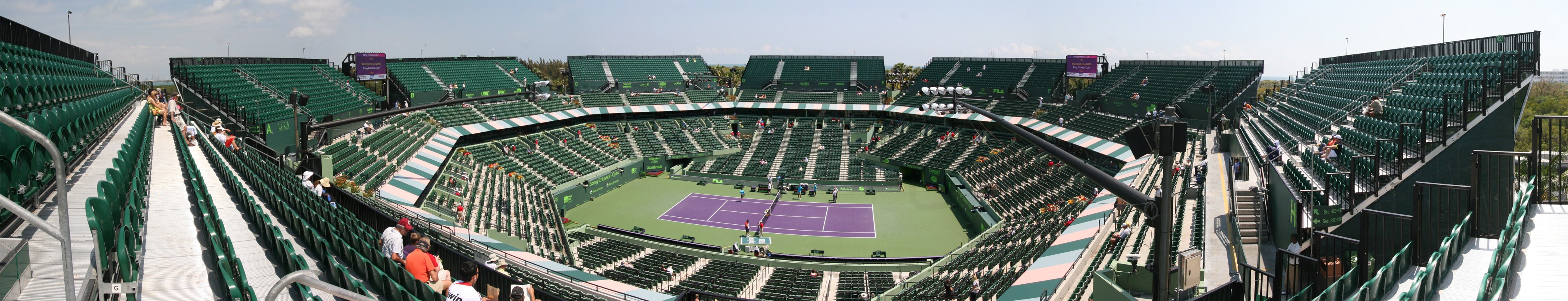 Panoramic view of a large tennis stadium with green seating sections and a purple hard court under partly cloudy skies.
