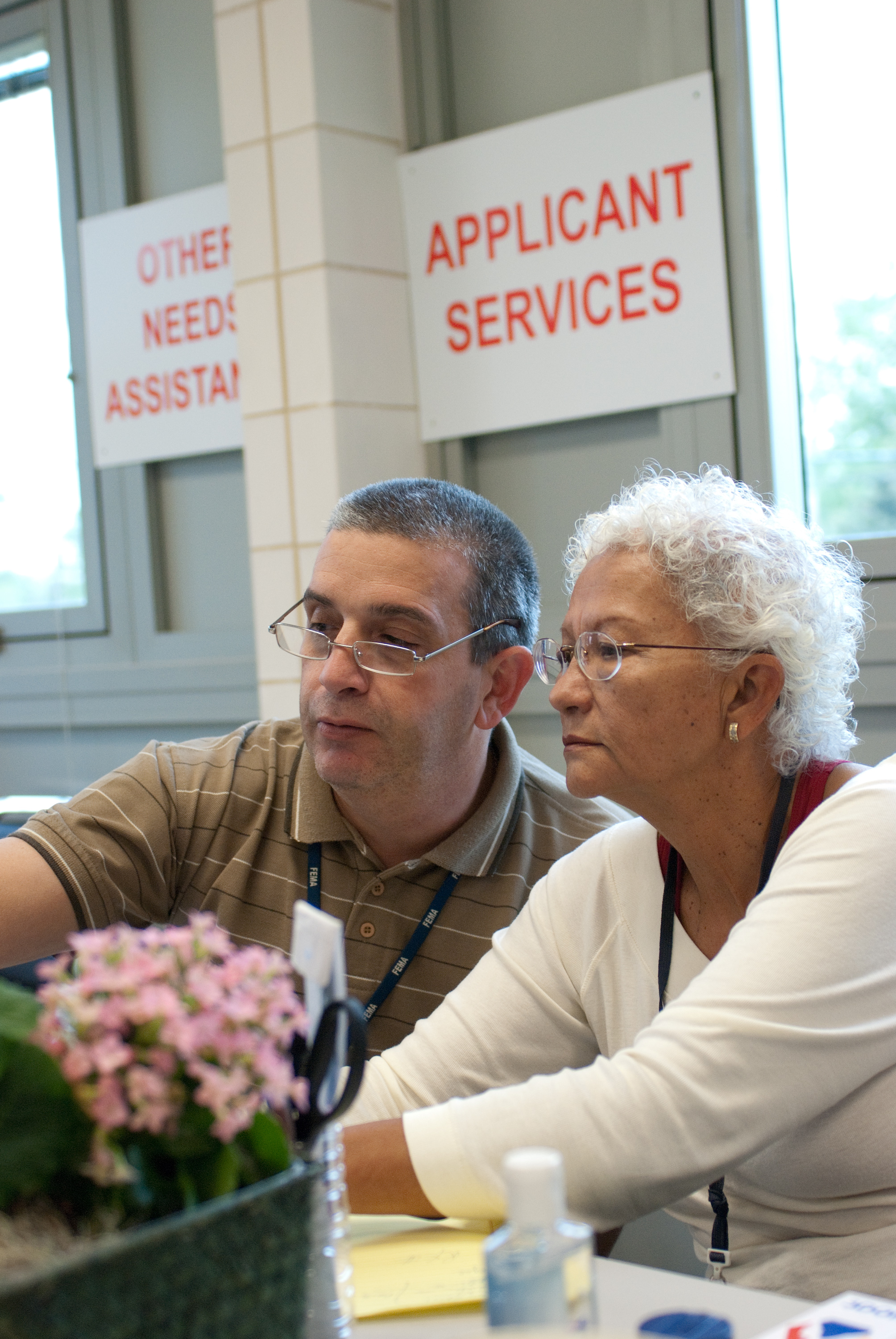 Two people sit at a desk inside a FEMA Disaster Recovery Center with assistance signs behind them.
