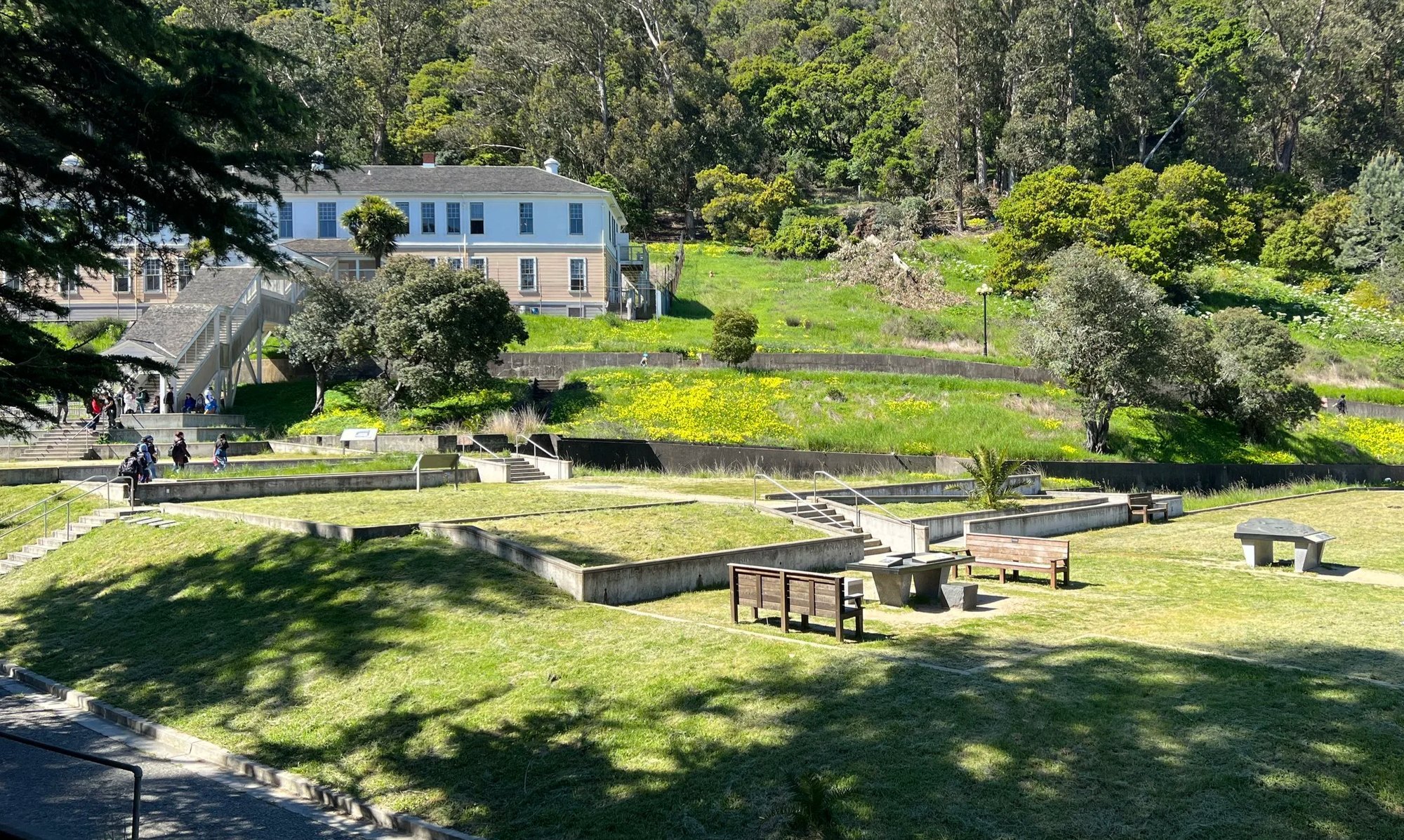 Terraced green hillside with benches and a white two-story historic building surrounded by trees and wildflowers.