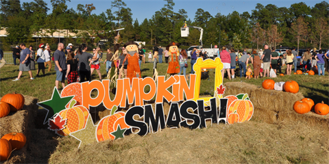 Colorful Pumpkin Smash sign surrounded by hay bales, orange pumpkins, and a crowd of families outdoors.