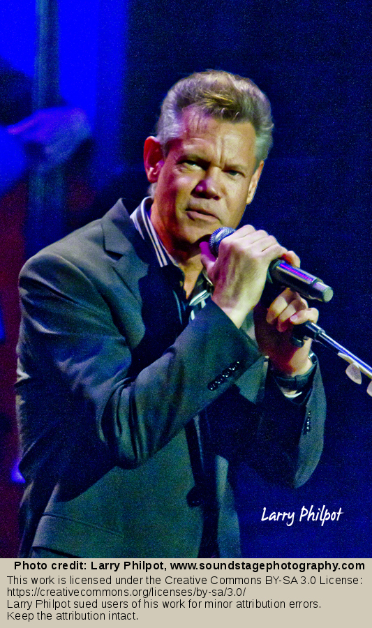 A man in a dark blazer sings into a microphone on a concert stage with blue lighting.