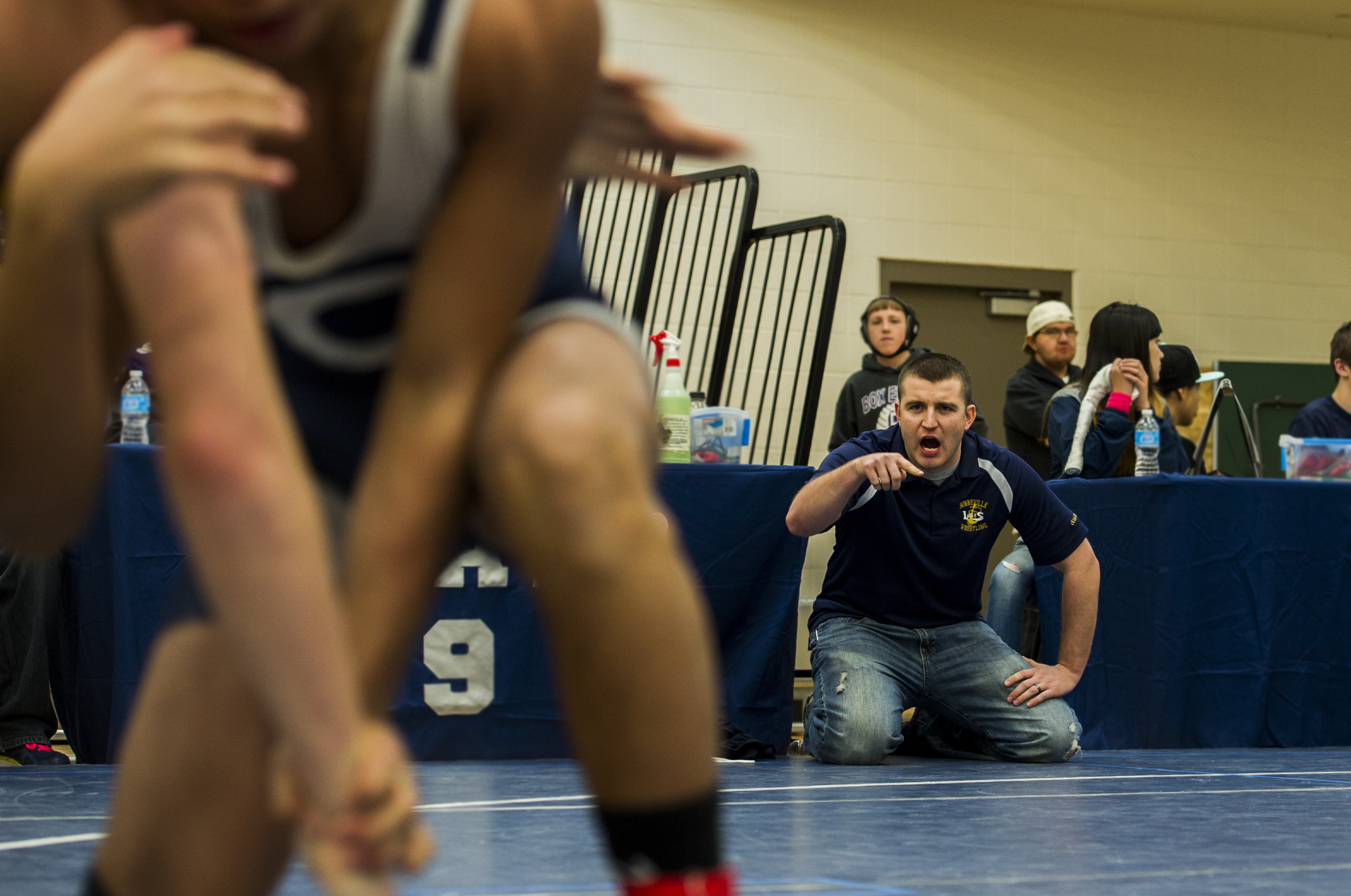 A coach kneels on a blue wrestling mat, pointing and shouting instructions.