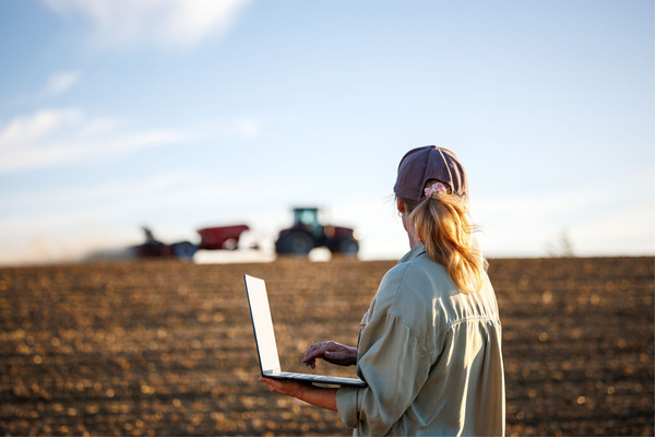 A person holds an open laptop in a tilled farm field with two tractors in the background.